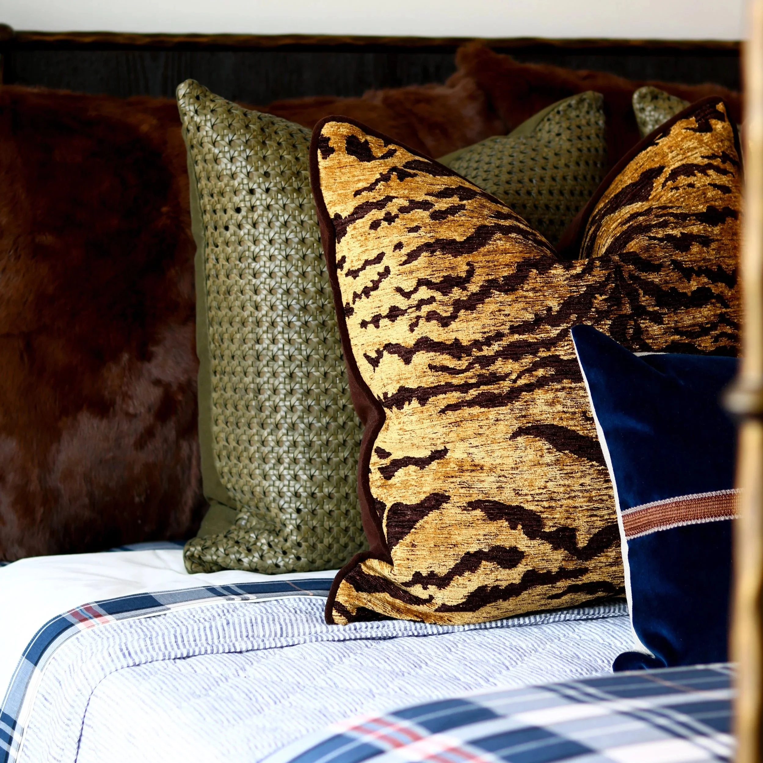 Close-up of decorative pillows on a bed, including patterned and animal print designs, with a striped bed sheet and a plaid blanket visible in the foreground.