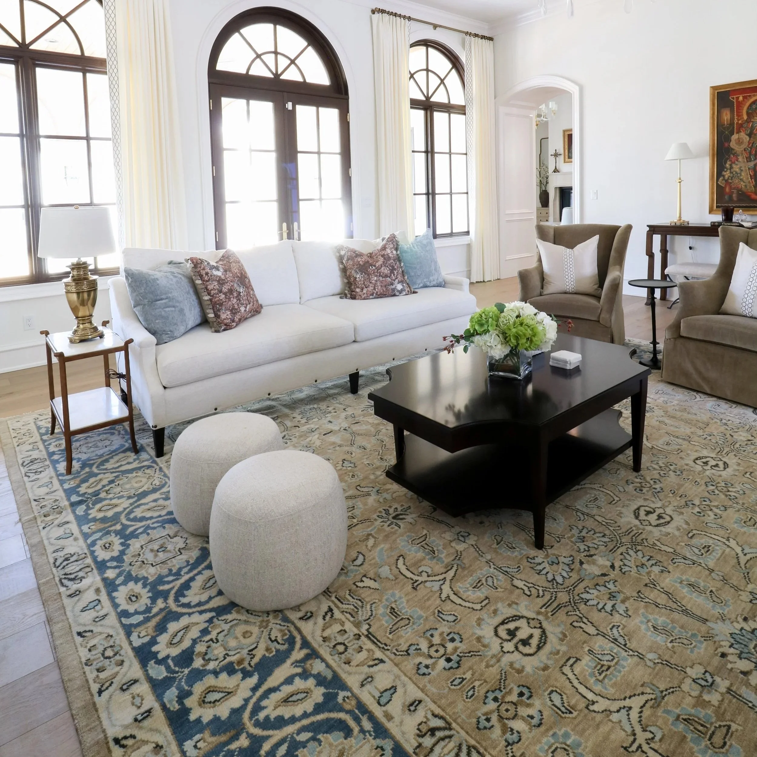 Living room with large arched windows, white walls, a white sofa with patterned pillows, two beige armchairs, a black coffee table with flowers, a side table with a lamp, and artwork on the walls.
