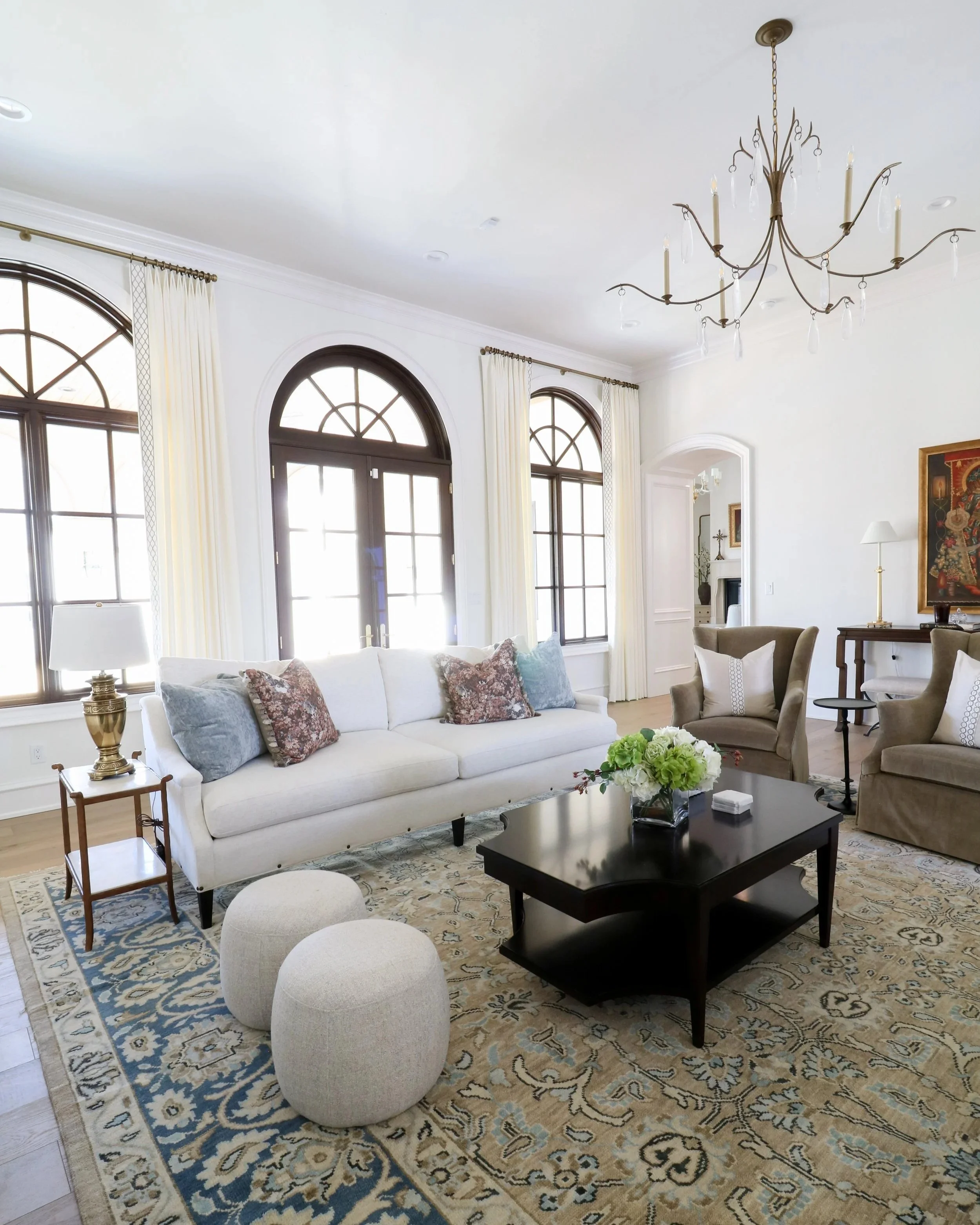 Bright living room with large arched windows, custom drapes, white walls, white sofa with patterned pillows, dark wood coffee table with flower arrangement, beige armchair, patterned area rug, chandelier, and side tables.