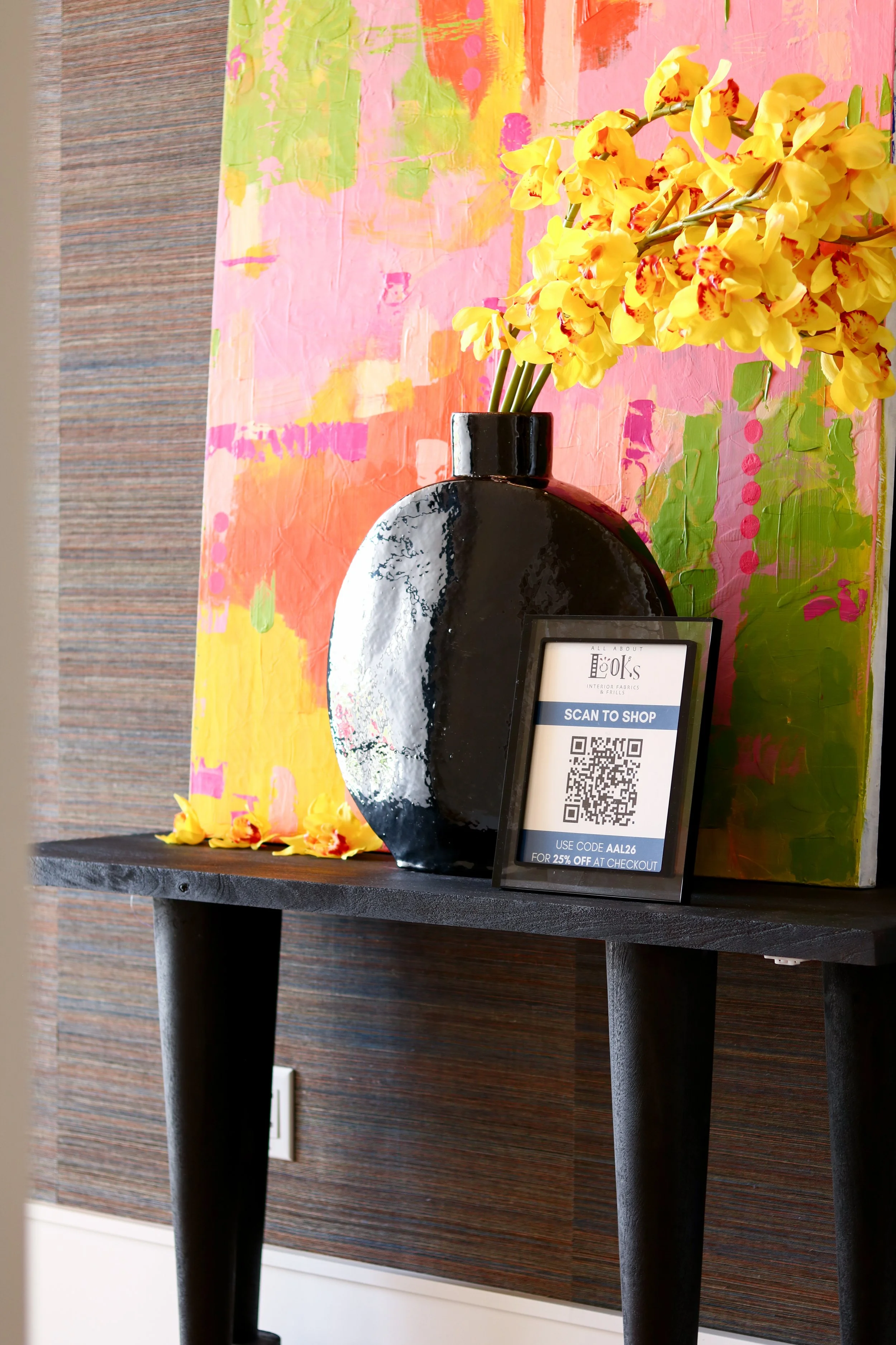 A black and white rounded vase with yellow orchids on a dark wooden table. A small sign with a QR code and text about a 25% discount stands in front of the vase. Behind, a colorful, abstract mural with pink, orange, green, and yellow shades covers th