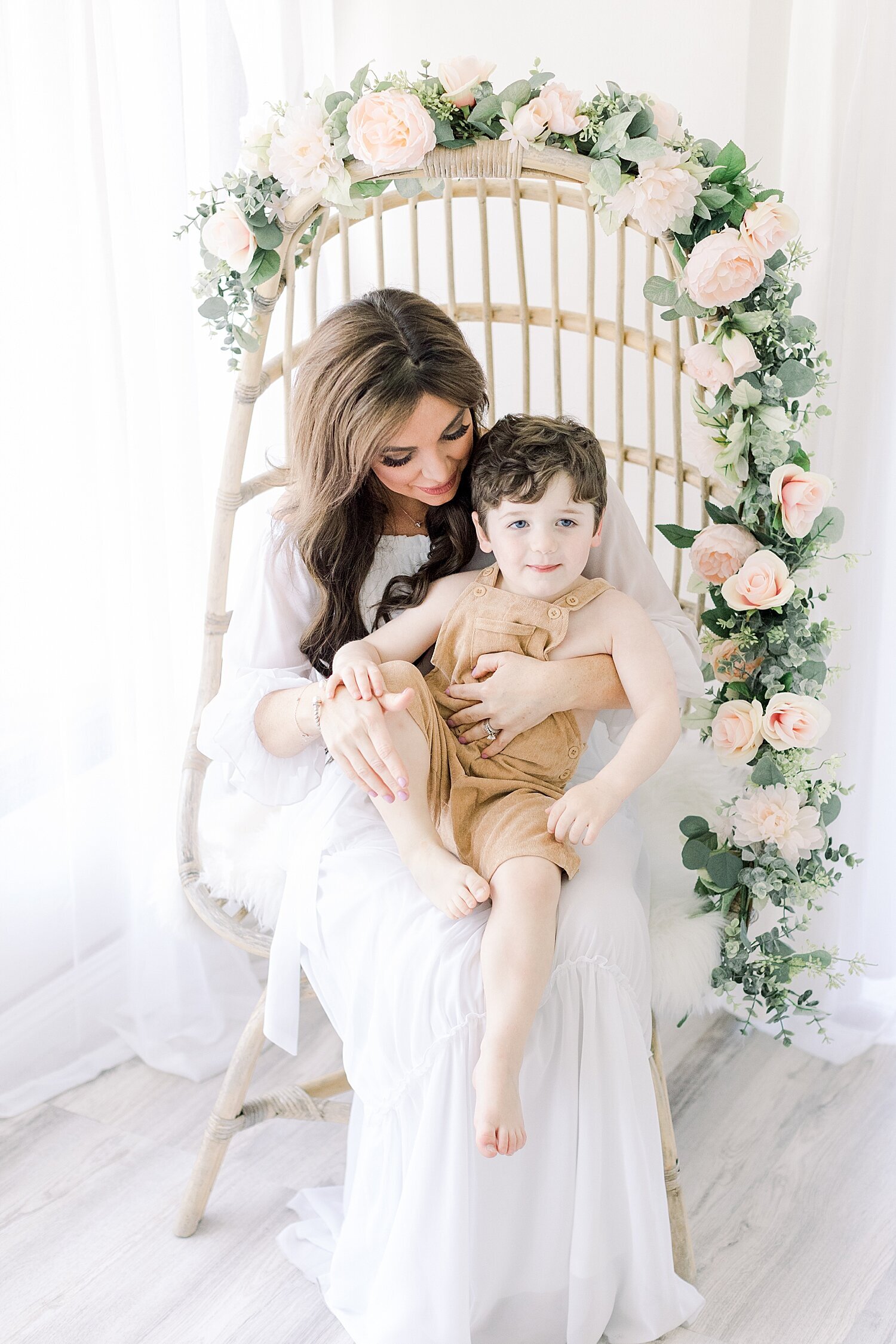 Mom sitting in rattan chair with floral garland holding her son. Photo by Ambre Williams Photography.