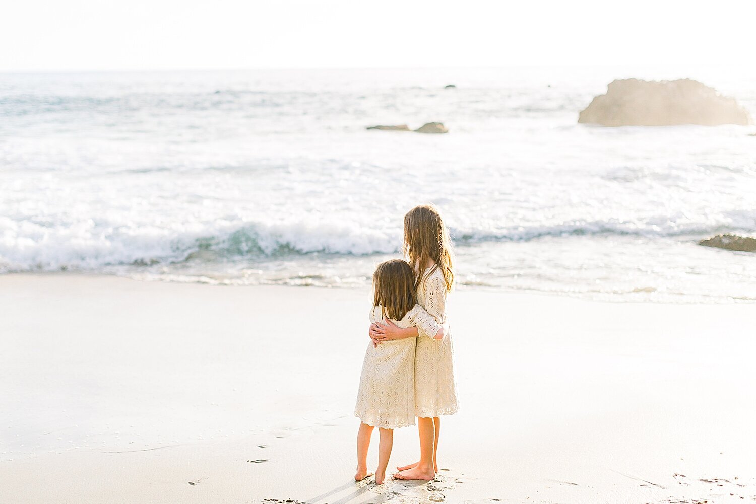 Two sisters hugging each other while standing in front of the waves during sunset in Newport Beach. Photos by Ambre Williams Photography.