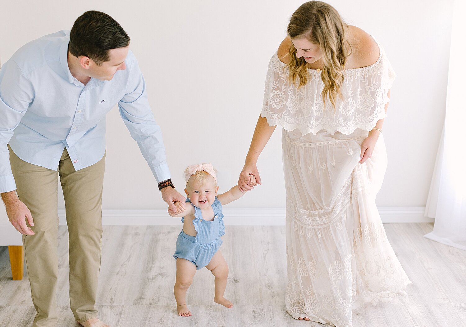 Mom and Dad holding their one year old daughters hand as she tries to walk. Photo by Ambre Williams Photography.