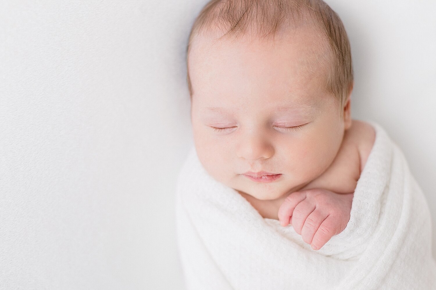 Baby boy swaddled in white with hands up by his chin. Photo by Ambre Williams Photography.