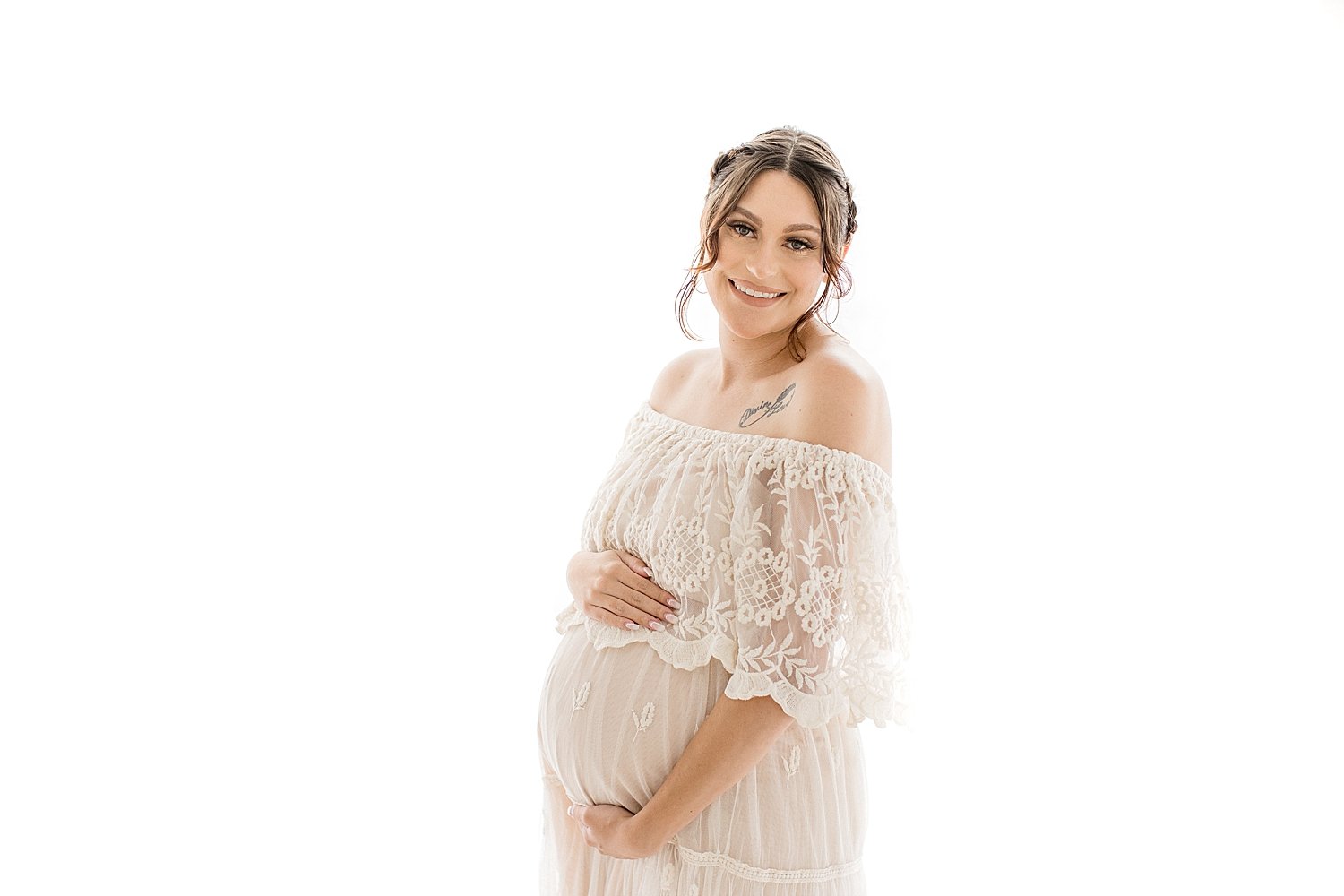Smiling pregnant mother in long sheer white dress holding her belly for maternity session in Newport Beach with photographer Ambre Williams