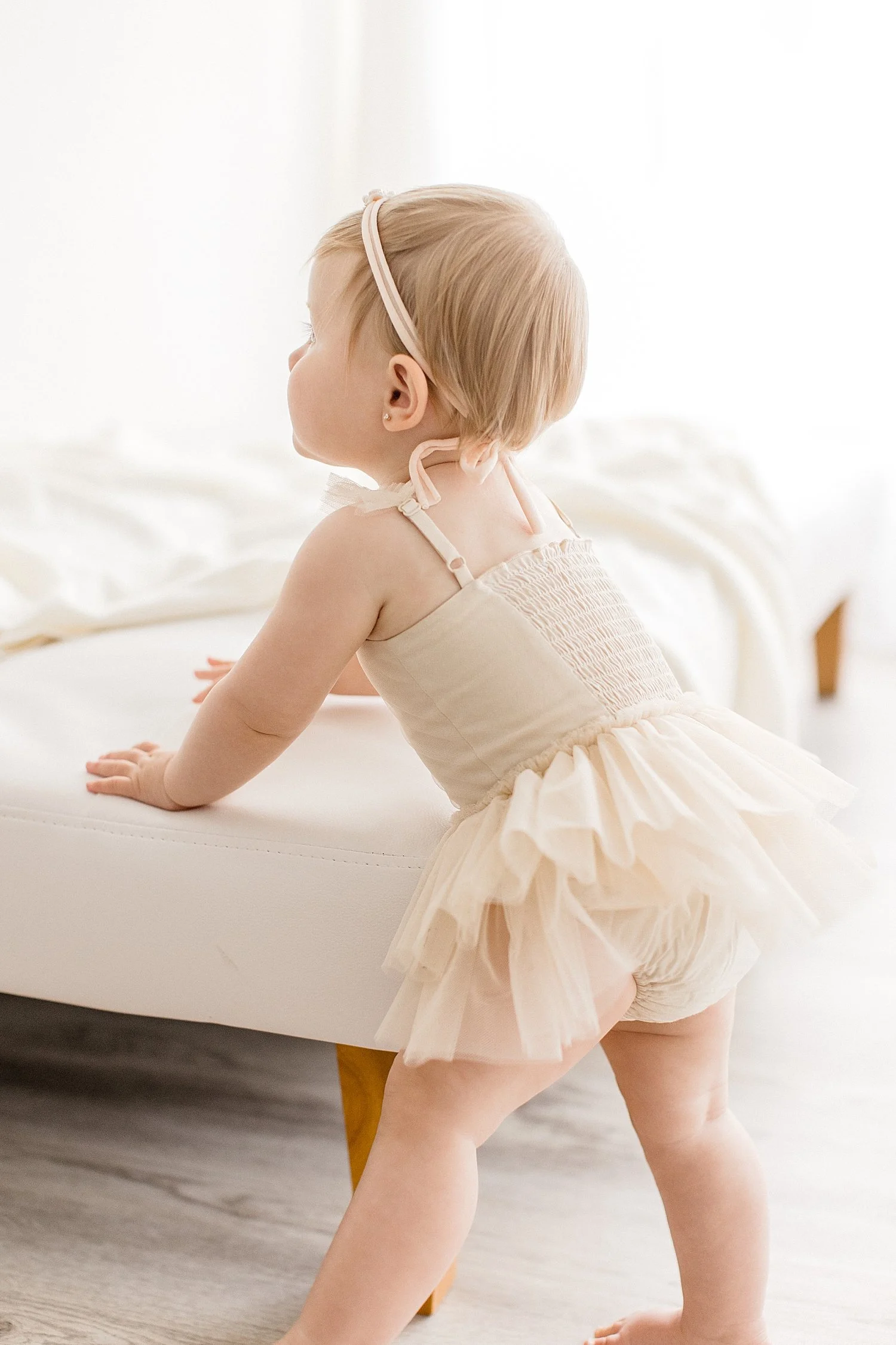 Baby girl wearing beige tulle leotard, looking away holding onto bench | Ambre Williams Photography