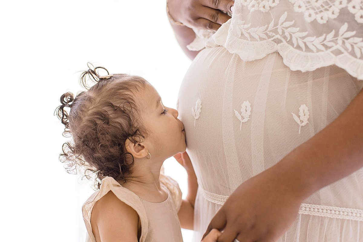 close-up of big sister kissing moms pregnant belly detail portrait with Ambre Williams Photography in her studio at Newport Beach