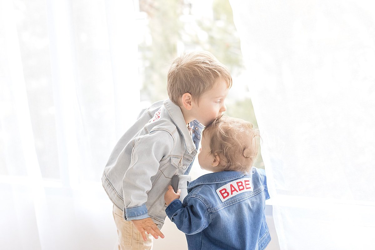 Big Brother giving Little Sister a kiss on the head in matching denim jackets during Newport Beach Studio Session with Ambre Williams Photography