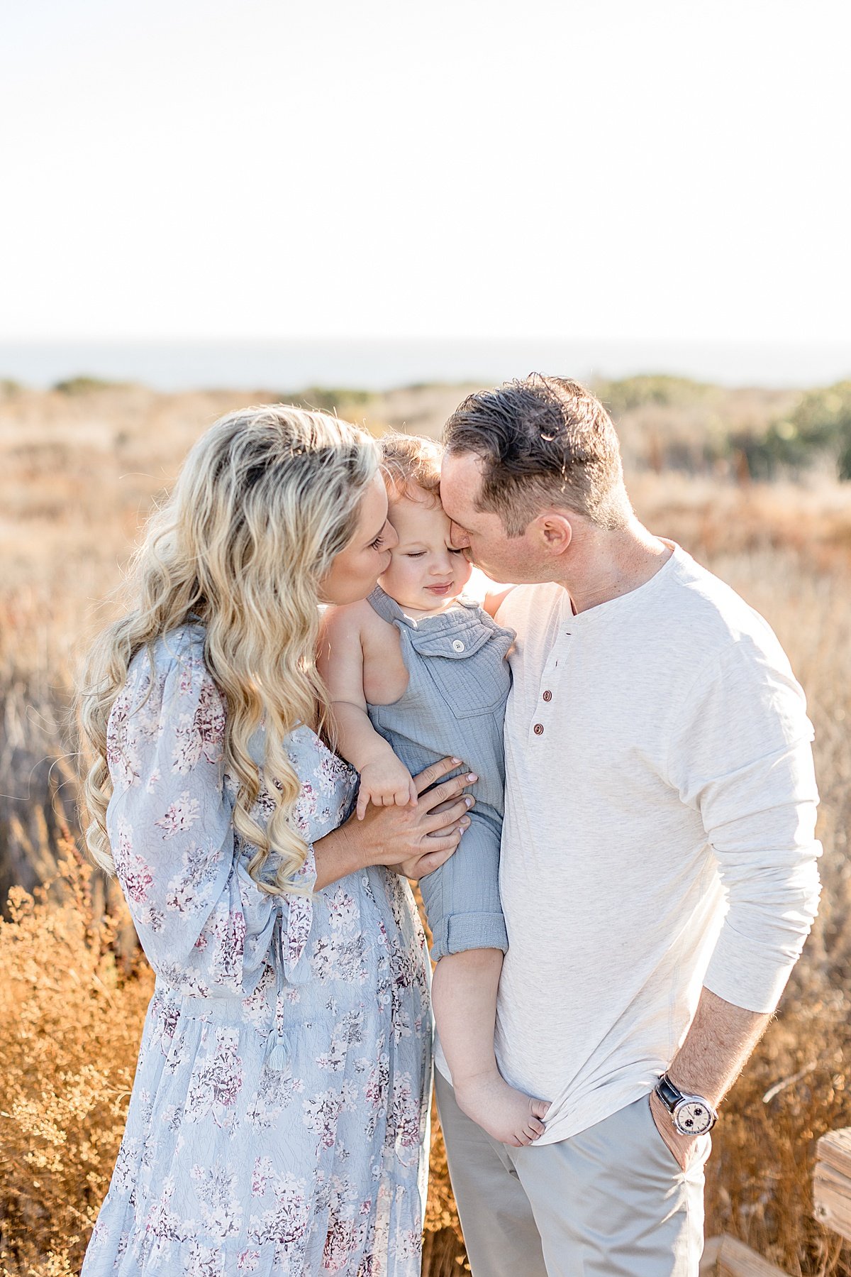 Outdoor family portrait, candid photography mom and dad kissing baby at beach with Ambre Williams Photography