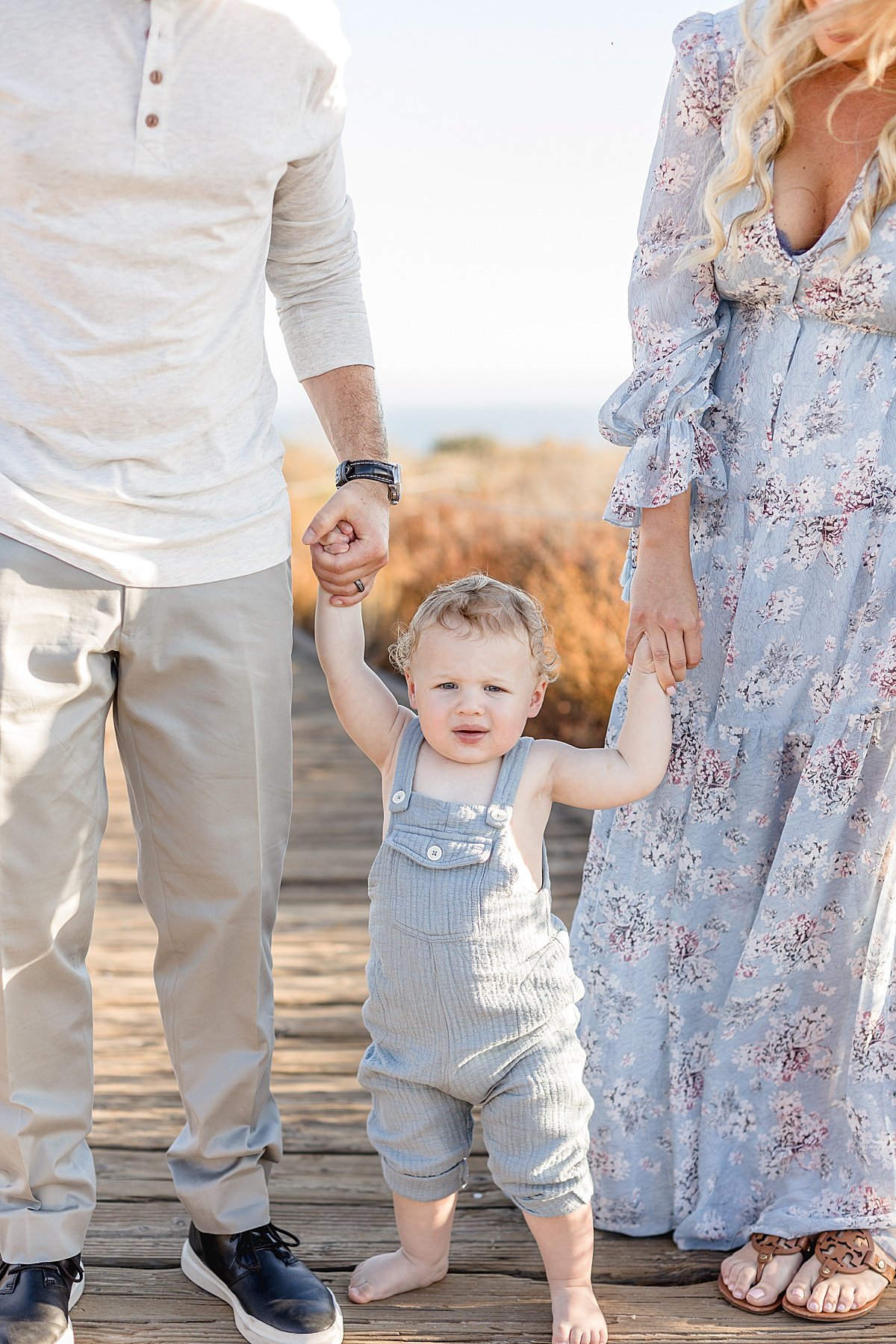 Mom and Dad holding happy standing babys hands during portrait session at sunny beach with Ambre Williams Photography in Newport Beach, California