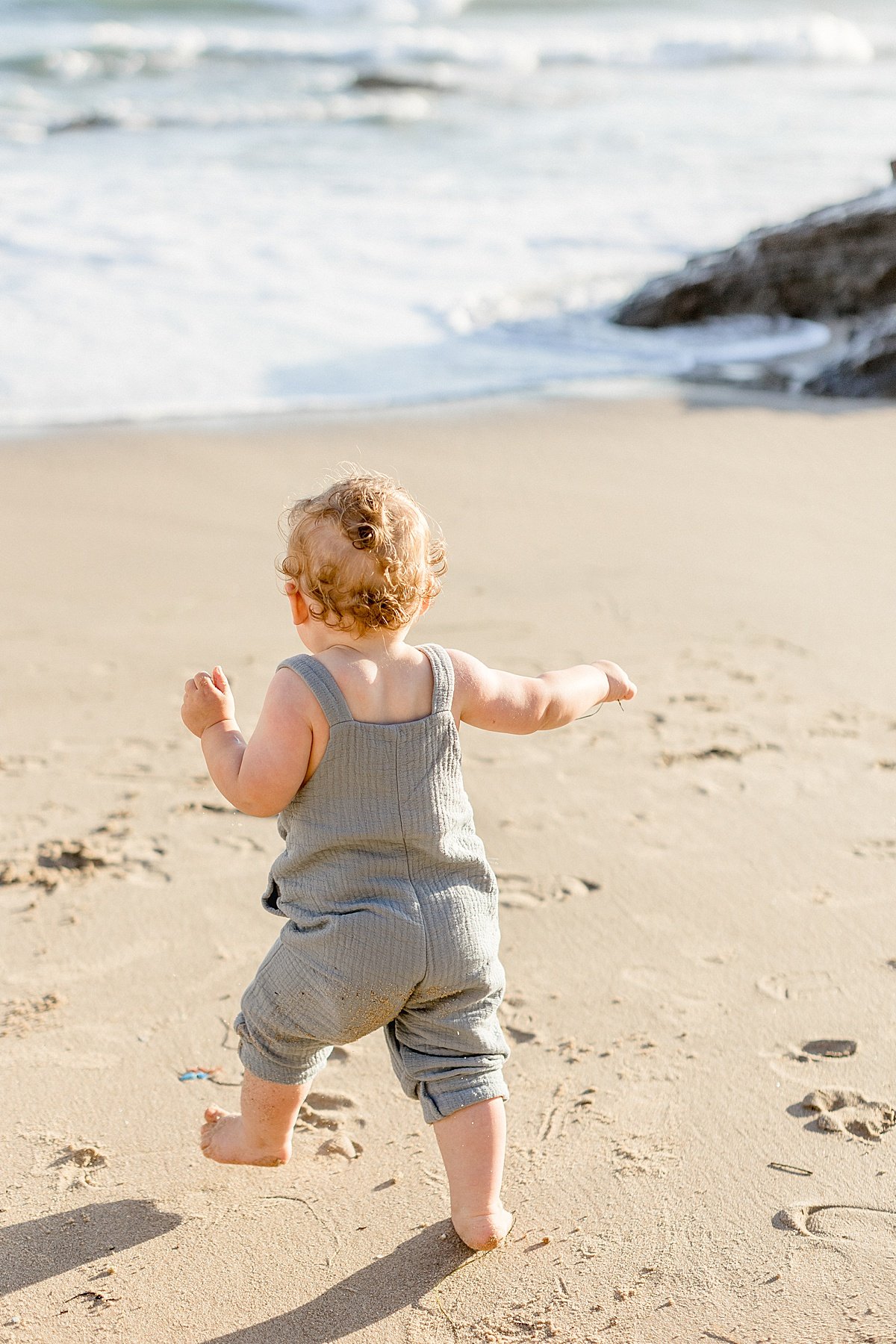 Candid portrait of happy baby boy in overalls running on beach during sunset | Ambre Williams Photography in Newport Beach, California