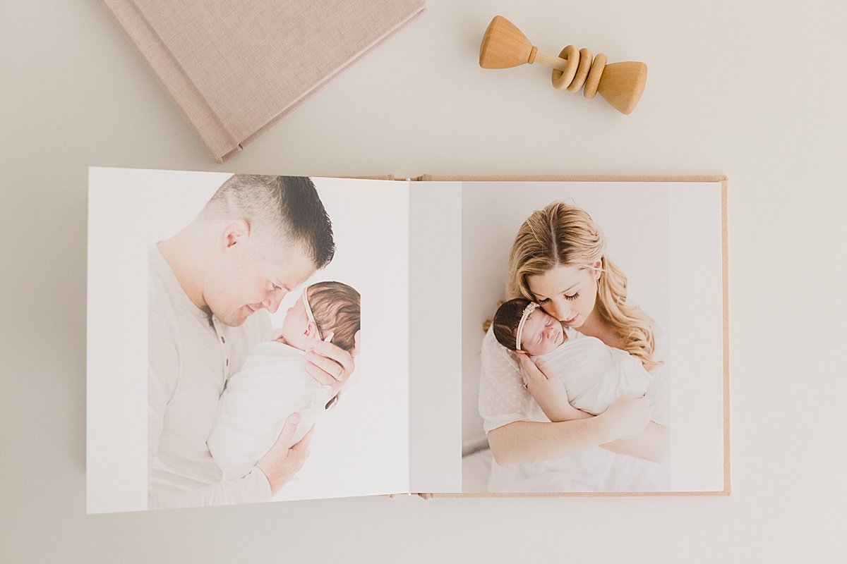 Flat Lay of open heirloom photo album of family holding their small newborn. Flat Lay has small baby toy. Photographed by Ambre Williams in Studio at Newport Beach