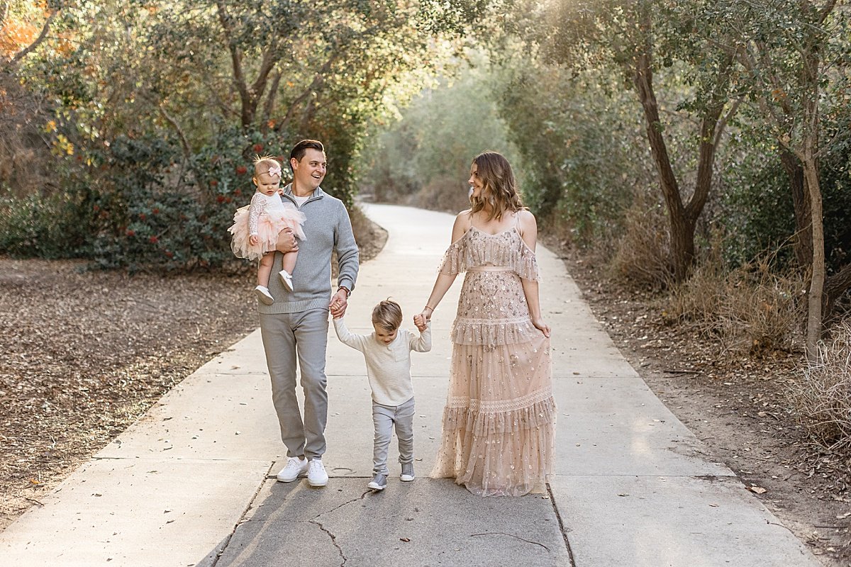 Happy family with two children smiling and walking together in Lake Forest road surrounded by trees during outdoor session with Ambre Williams Photography