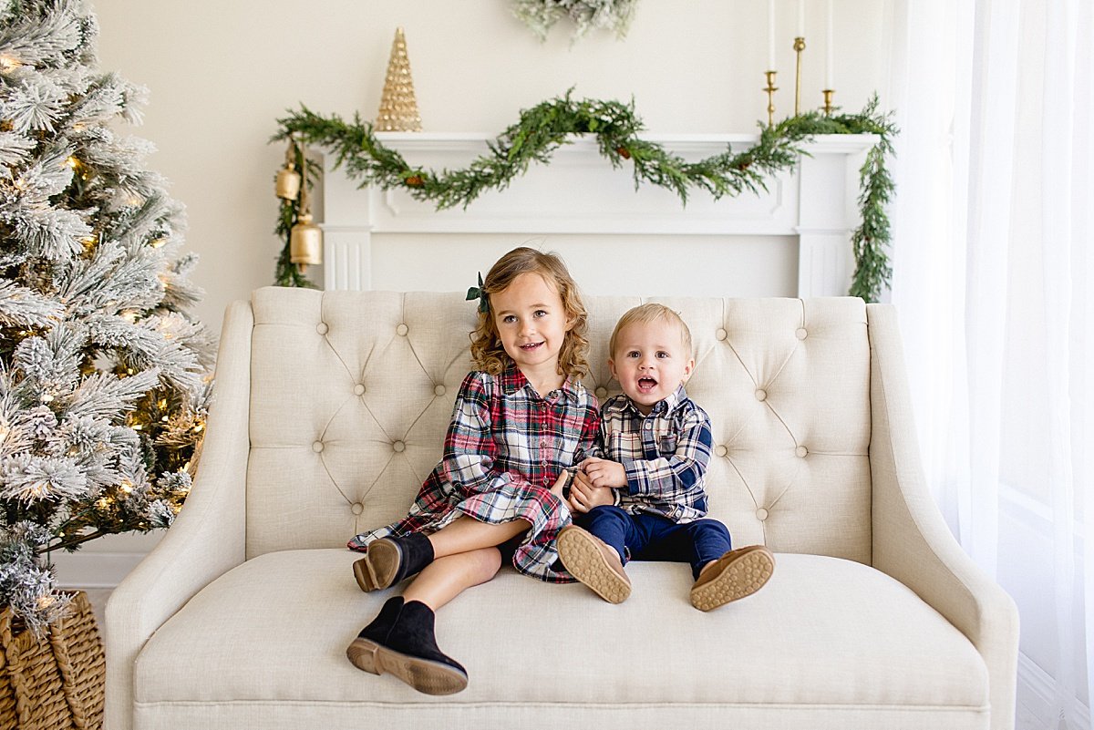 Two children sitting on white couch during santa magical experience in Ambre Williams Photography studio in Newport Beach