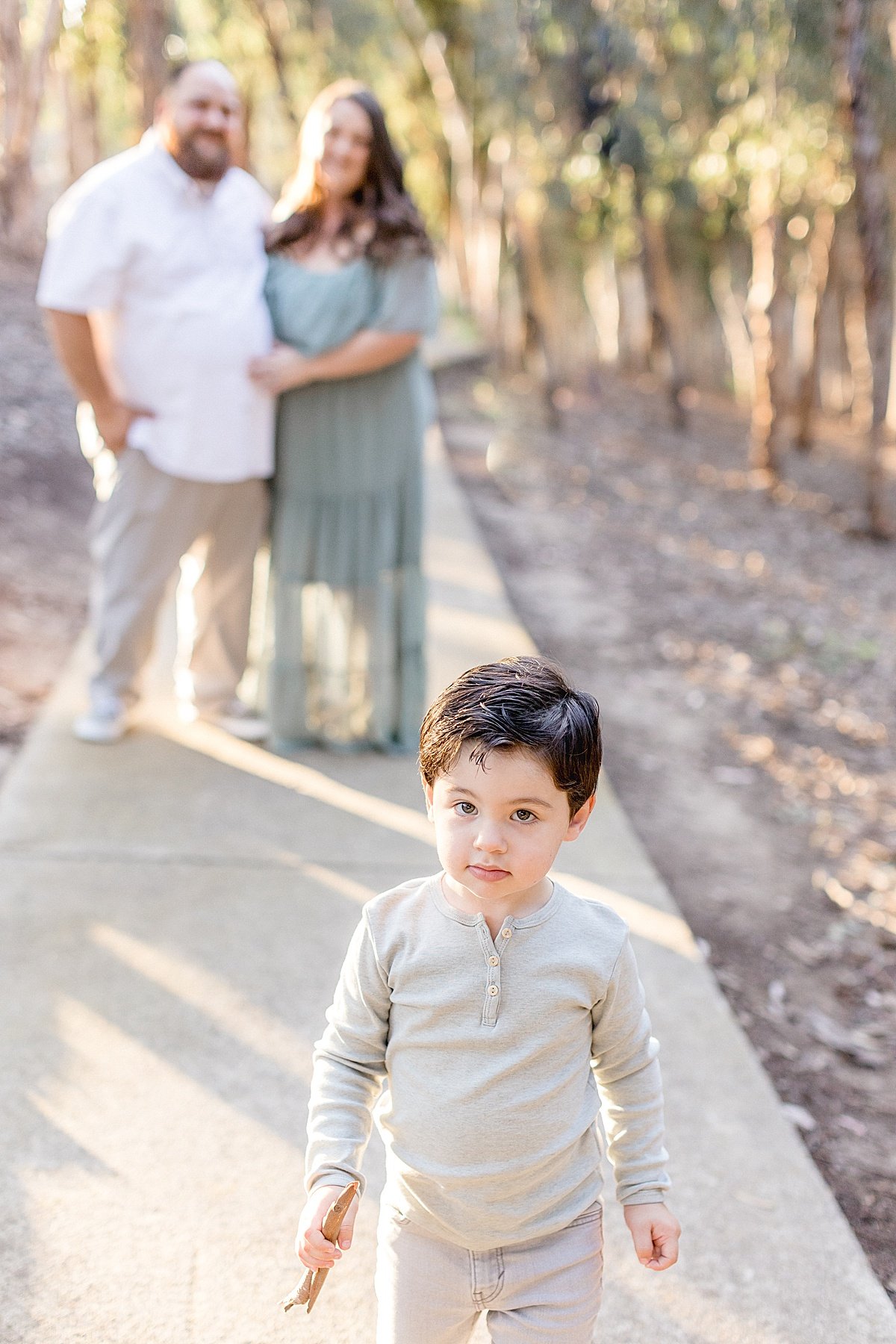 Mom and dad smiling together as son walks away during outdoor portrait session in Lake Forest with Ambre Williams Photography