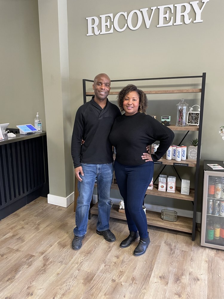 Owners Giovanni and Sharquana are standing in front of shelves stocked with CBD products inside their store which is painted in a calm neutral color. The word "Recovery" is on the wall over their heads.