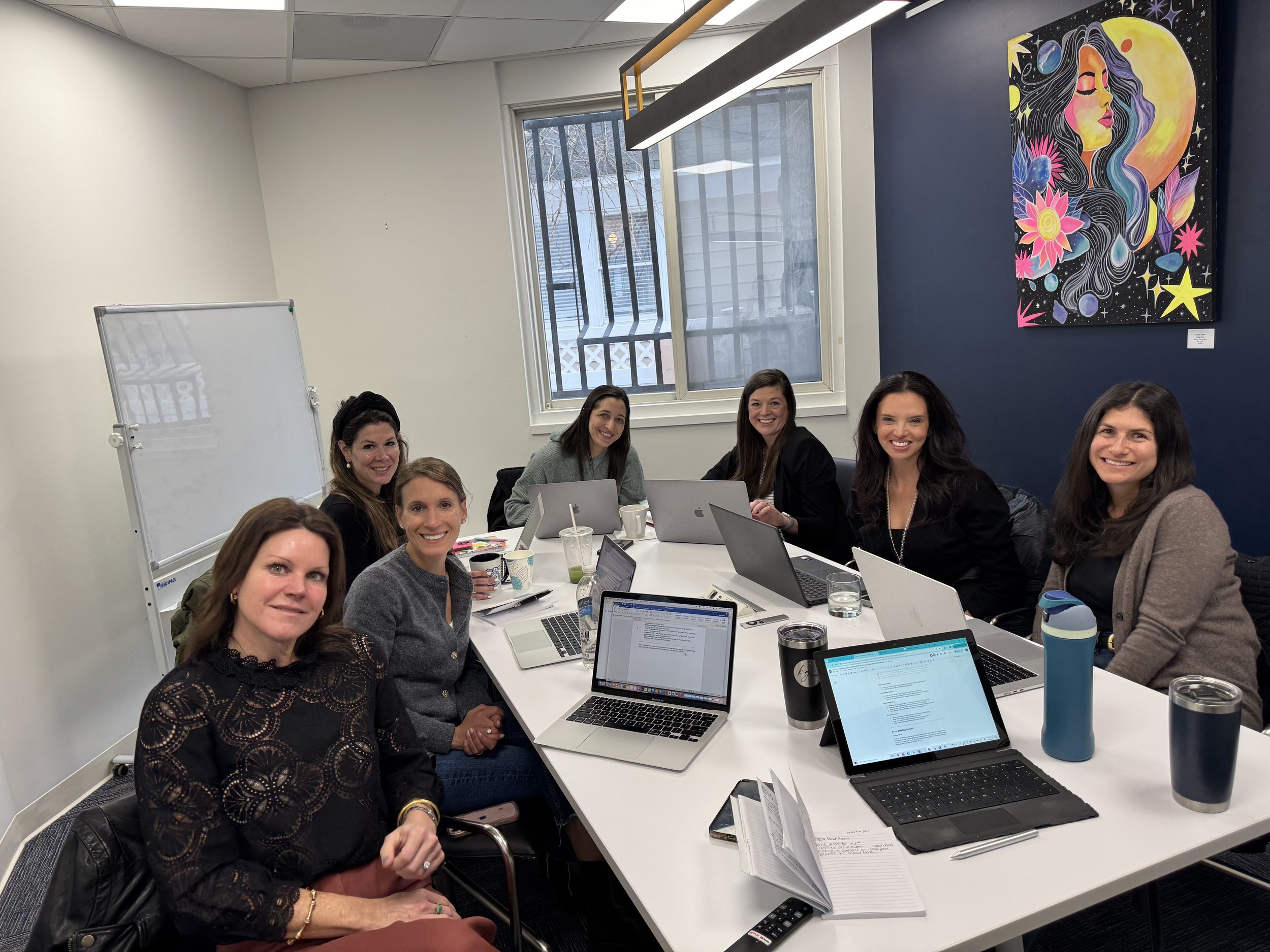 A group of nine women sitting around a conference table in a modern office with laptops, notebooks, and drinks, smiling at the camera. The room has a whiteboard, a large window, and a colorful mural of a woman's face with celestial and floral elements on the dark blue wall.