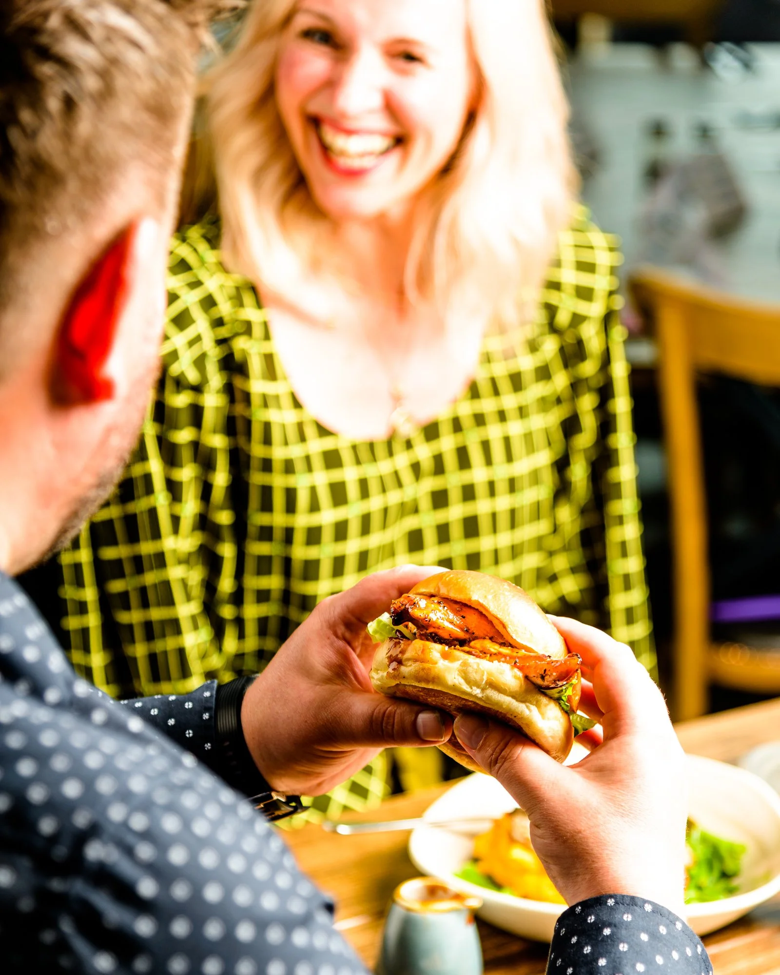 A woman with blonde hair smiling in the background as a person in the foreground holds a burger with lettuce, tomato, and grilled chicken bun. The scene is in a cozy restaurant or cafe setting.