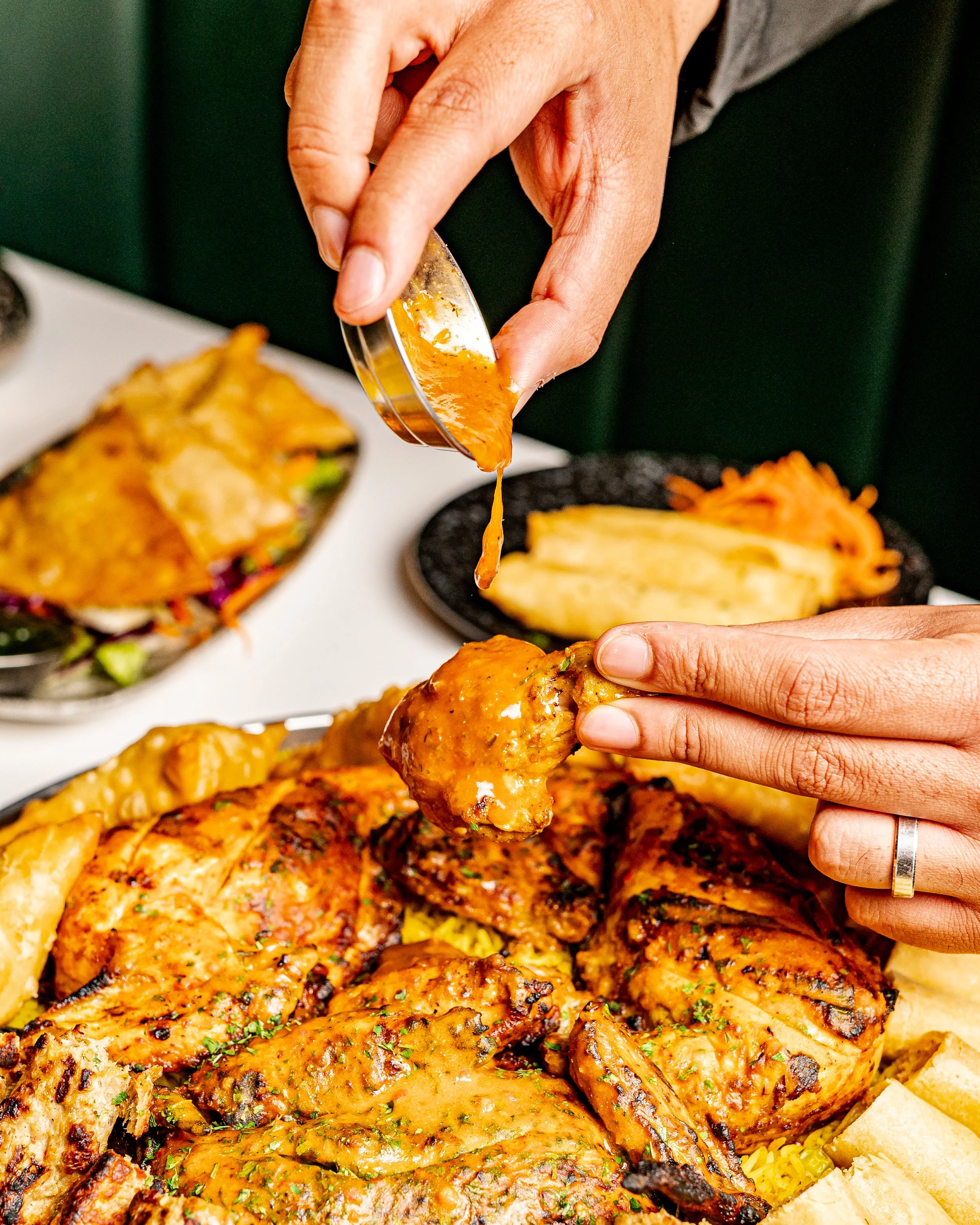 A person is holding a piece of grilled chicken with a serving of sauce above a platter of assorted grilled chicken, fries, and pasta.