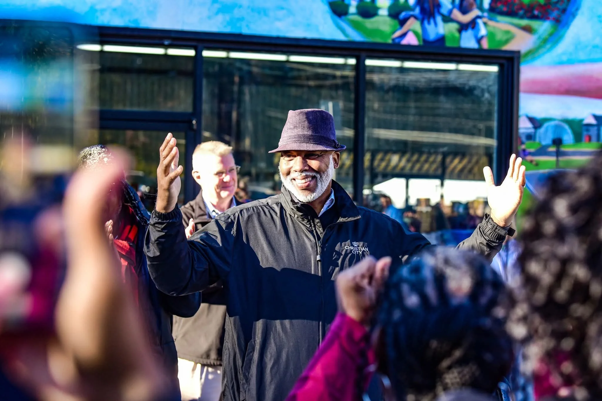 A group of people standing in front of a colorful mural on a building, participating in a ribbon-cutting ceremony, with a crowd gathered around and some trees in the background.