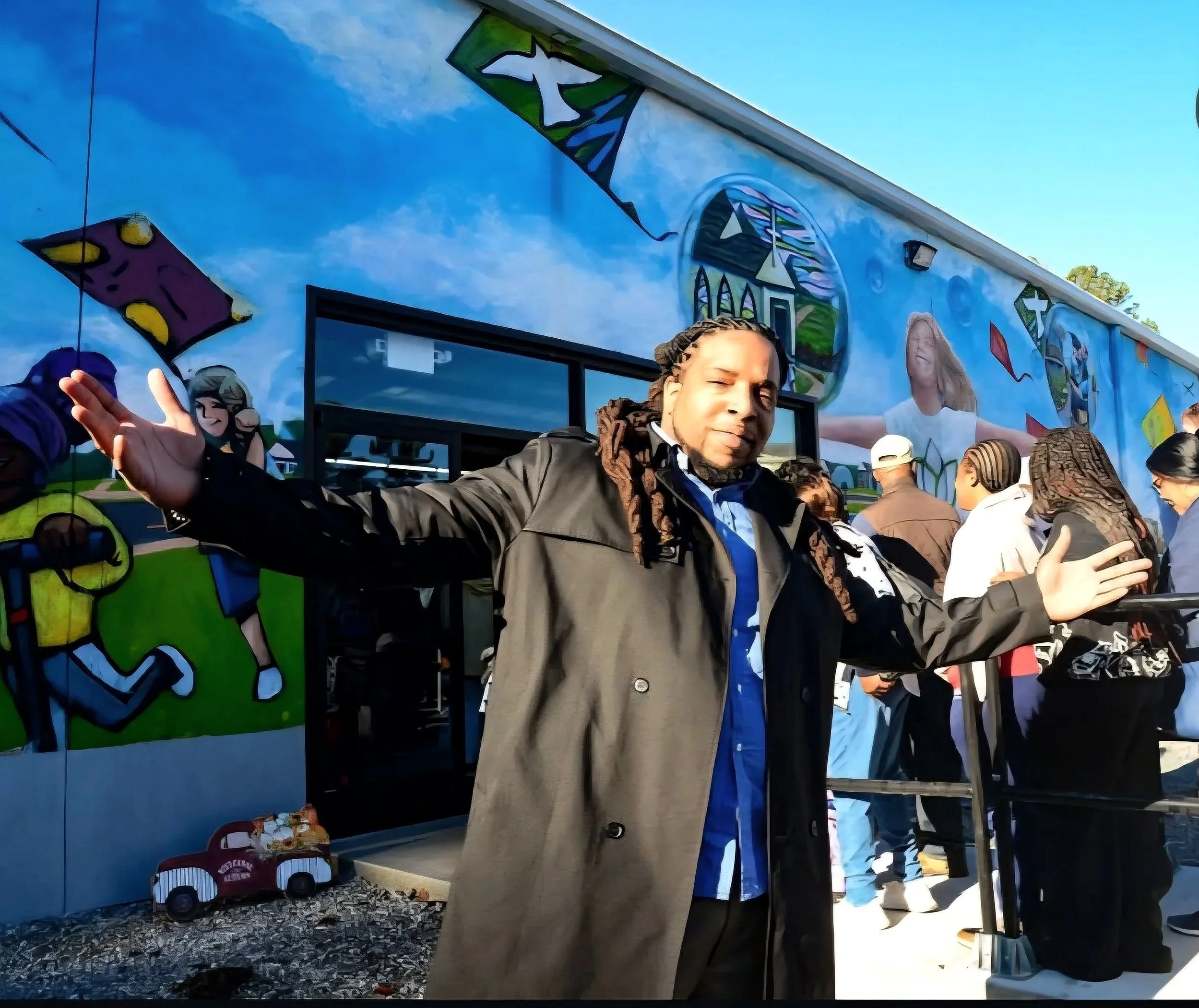 A person with dreadlocks wearing a black jacket stands in front of a colorful mural, with arms outstretched, amidst a group of people in line.