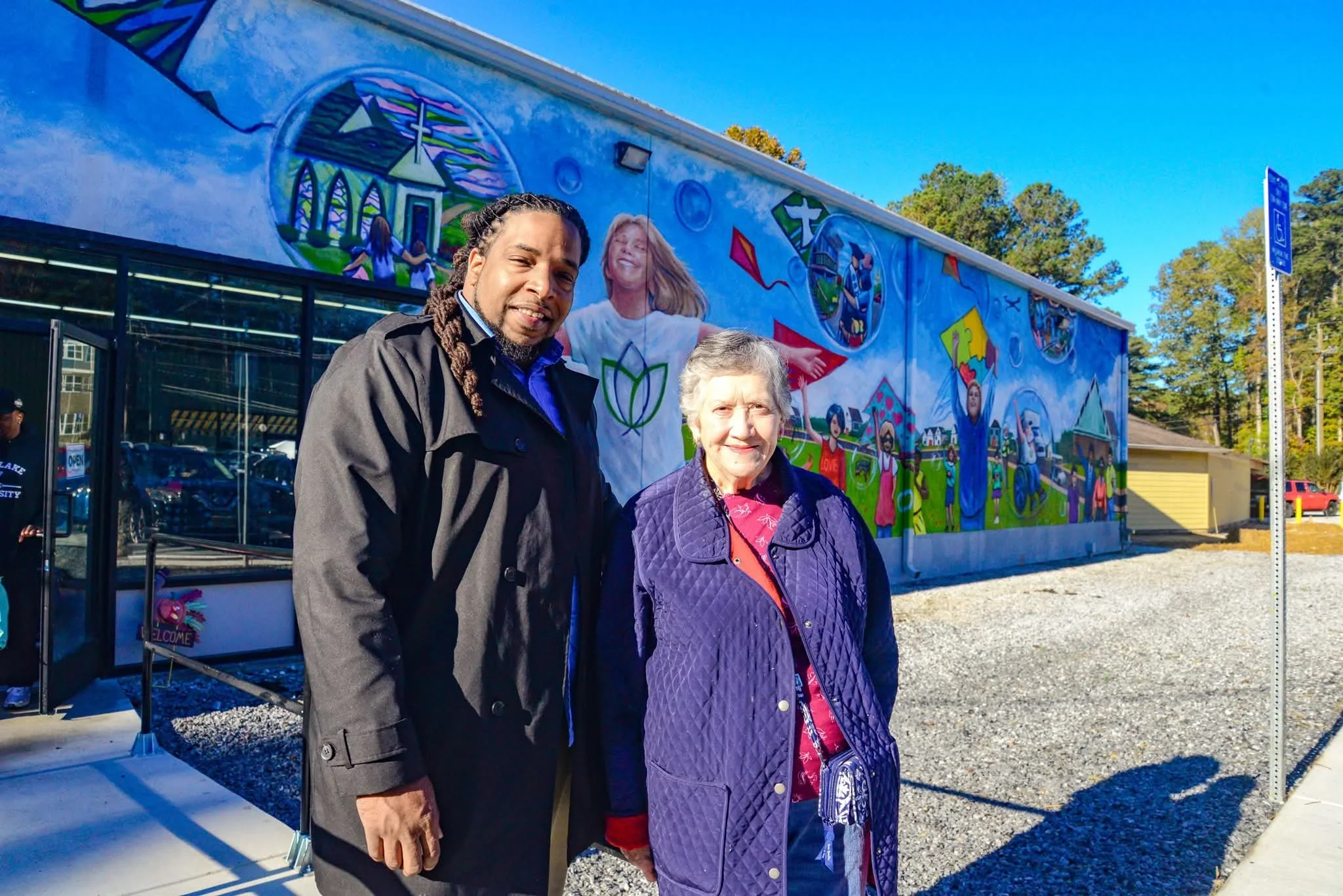 Two people standing in front of a colorful mural on a building, with trees and a clear blue sky in the background.