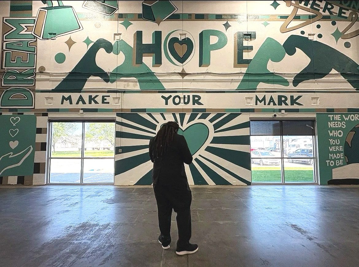 A woman standing inside a building with a large mural on the wall that reads 'MAKE YOUR MARK' and 'HOPE' with two hands holding a heart and a few stars, and a smaller text reading 'THE WORLD NEEDS WHO YOU WERE MADE TO BE.' The mural features teal, black, and white colors with windows showing parking lot and trees outside.