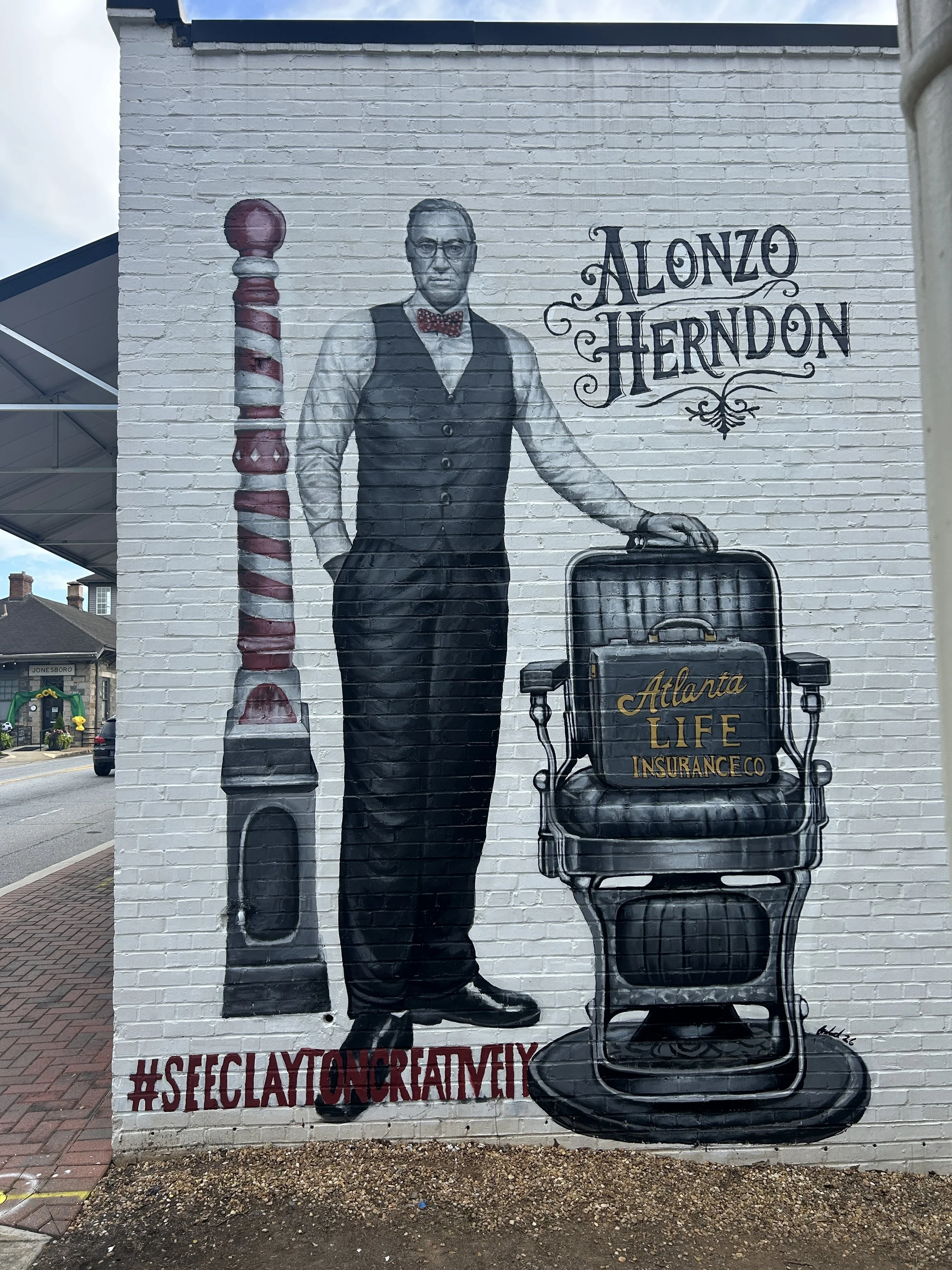 A mural of a man dressed in vintage attire, including a vest, bow tie, and glasses, standing next to a barber pole and a barber chair with a large suitcase. The mural includes text reading 'Alonzo Herndon' and 'Atlanta Life Insurance Co.' The hashtag '#SCECLAYONCRAFEMY' is painted at the bottom.