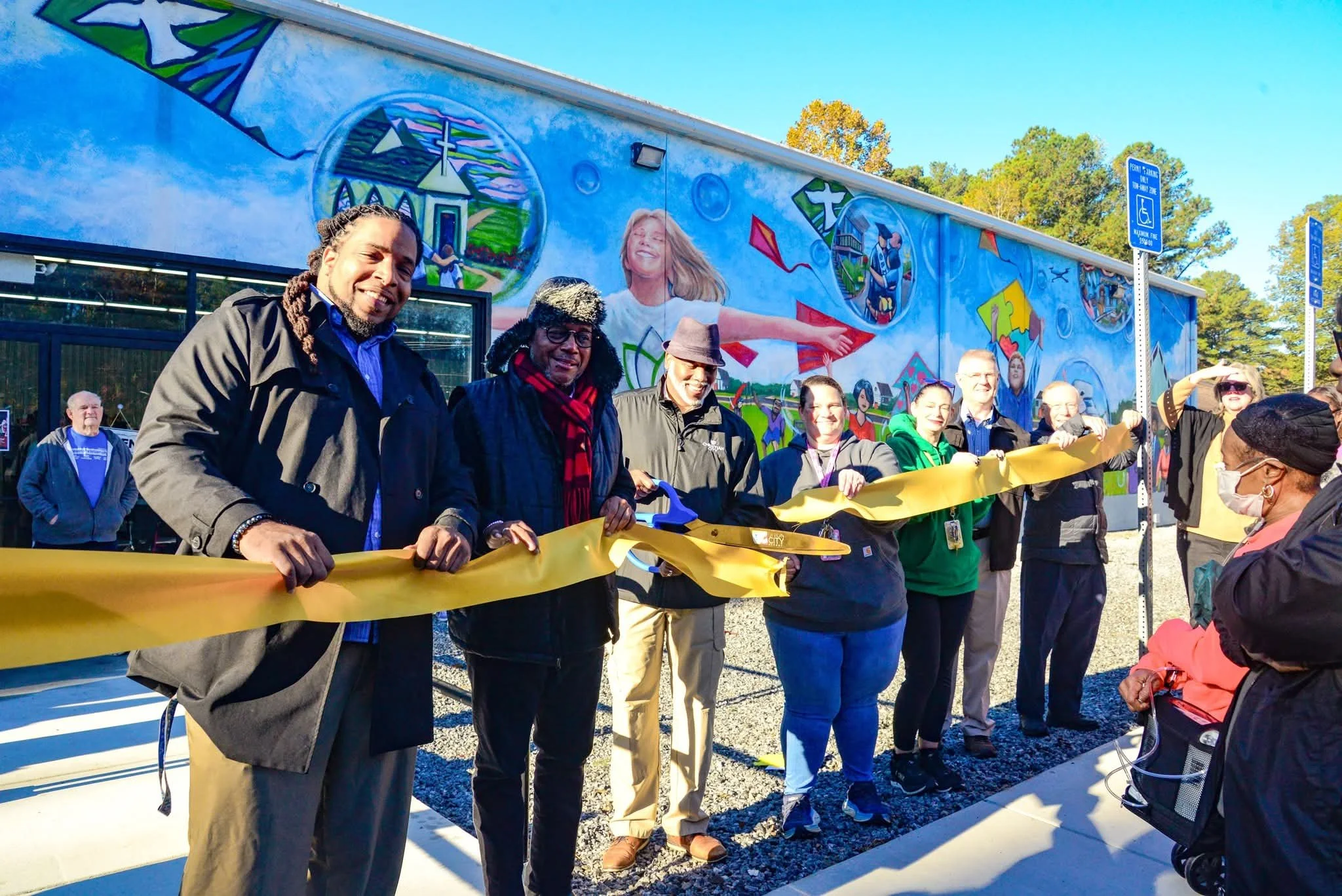 Group of people at a ribbon-cutting ceremony outdoors in front of a colorful mural on a building wall, with some individuals holding a yellow ribbon and scissors, and a person on the right wearing a face mask.