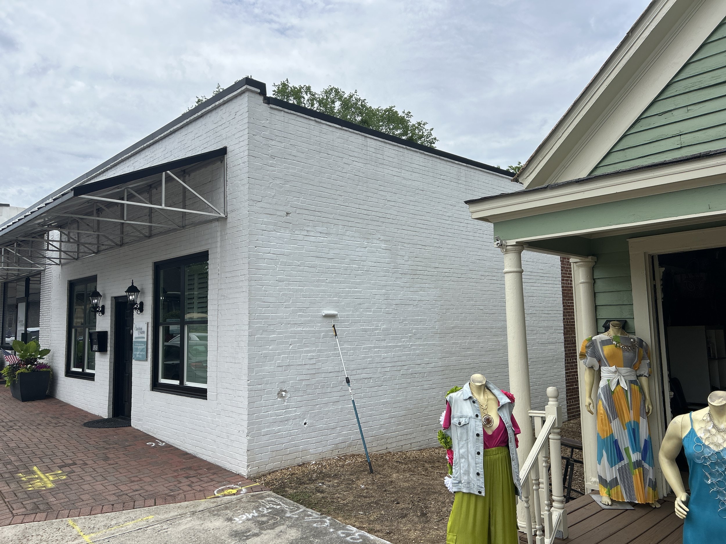 Exterior of a white brick building with black trim, large windows, and outdoor lighting. To the right, part of a porch with clothing mannequins dressed in colorful outfits, one with a striped dress and another with a denim vest and bright skirt. A small tree is visible behind the building, and the sky is partly cloudy.