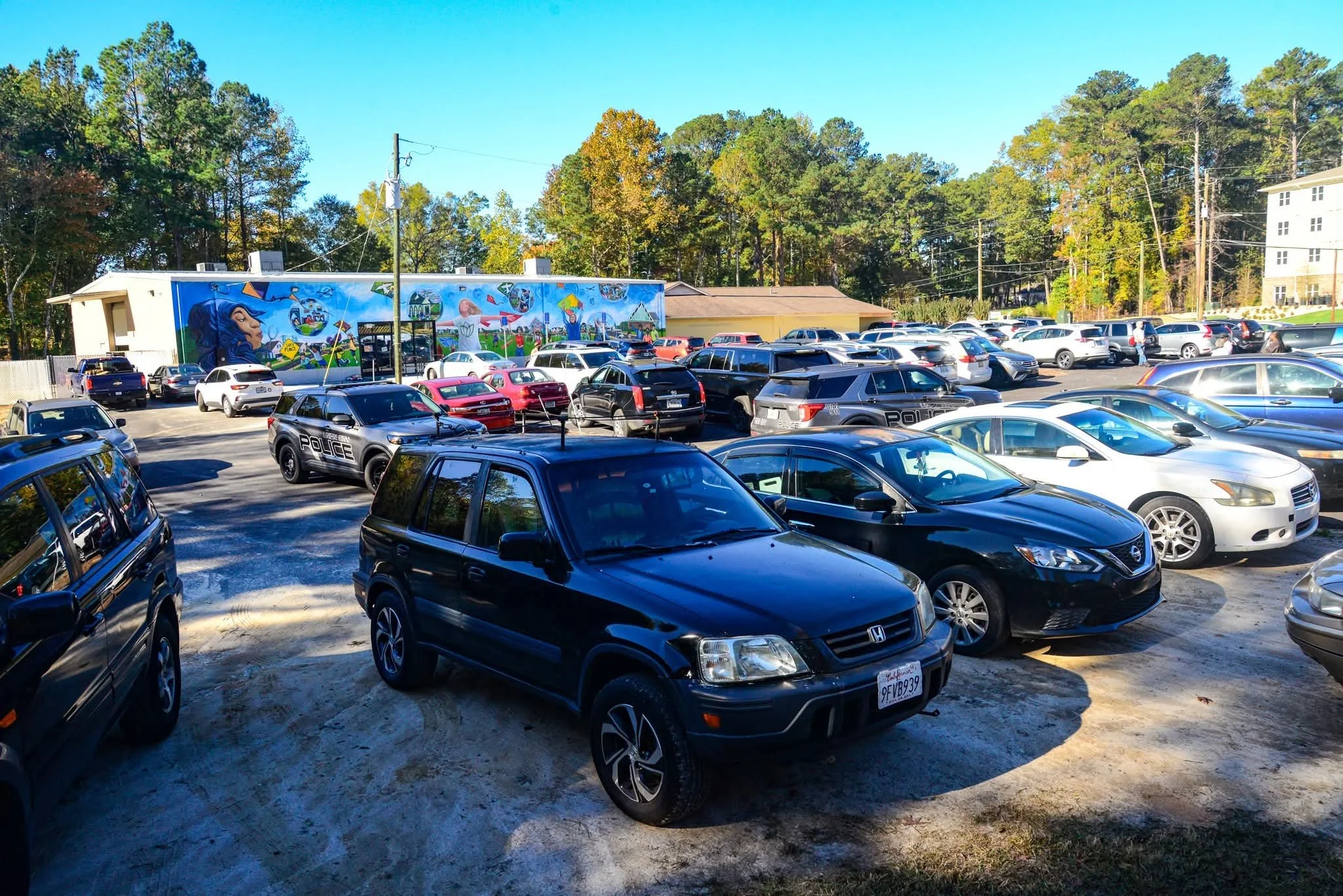 A crowded parking lot filled with various cars, including police vehicles, with a colorful mural on a building in the background and trees beyond.