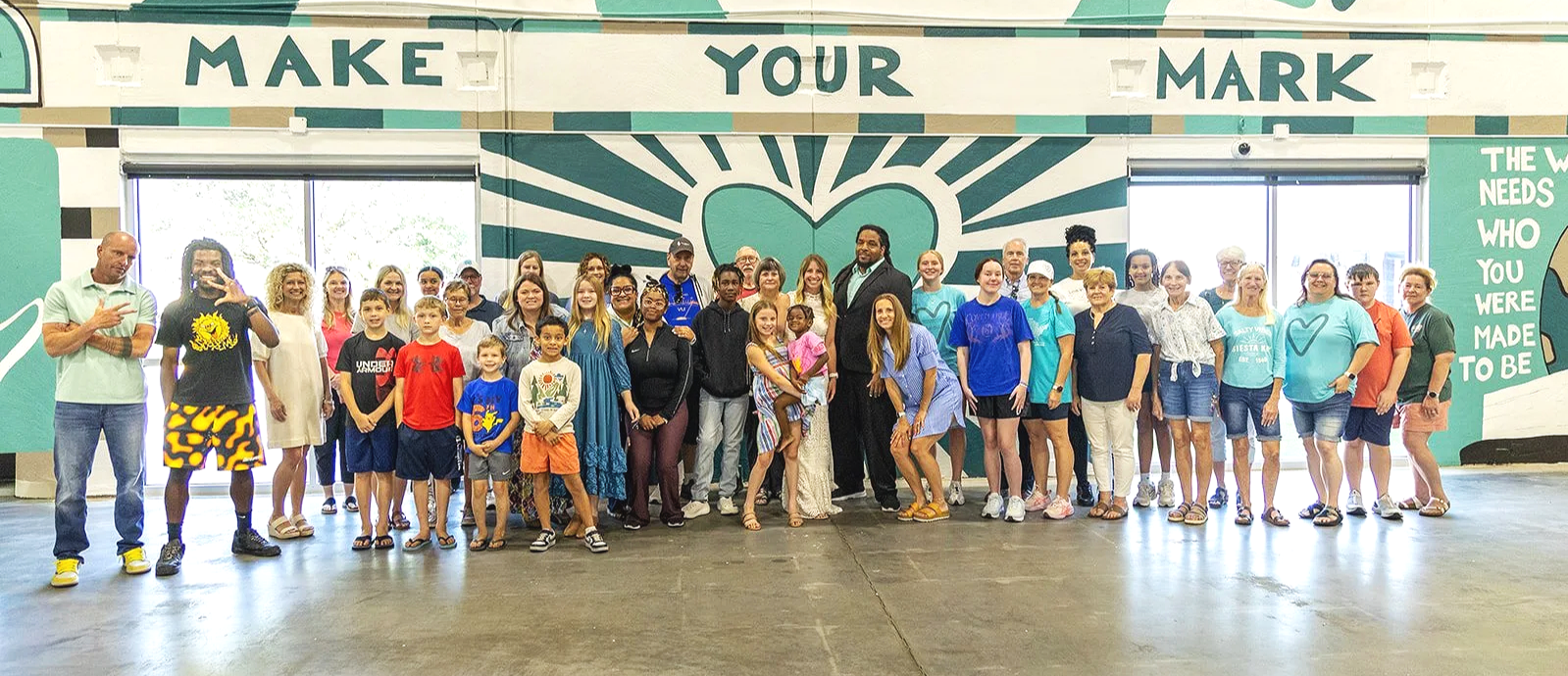 A large group of people, including children and adults, standing inside a building with a mural that reads "Make Your Mark" and a big heart design on the wall behind them, posing for a group photo.