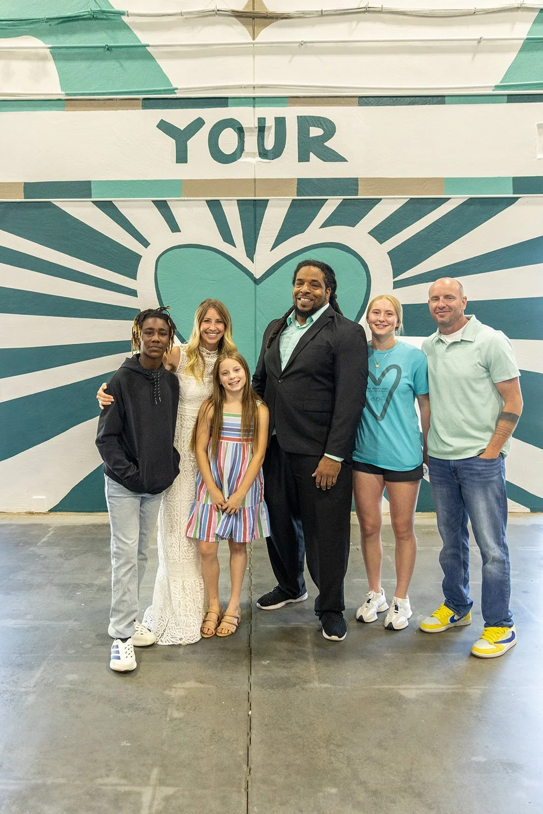 A group of six people, three young women, two young girls, and one man in a suit, standing in front of a mural with a heart design and the words "YOUR" at the top. The background is painted with teal, white, and beige colors, and the group is smiling.