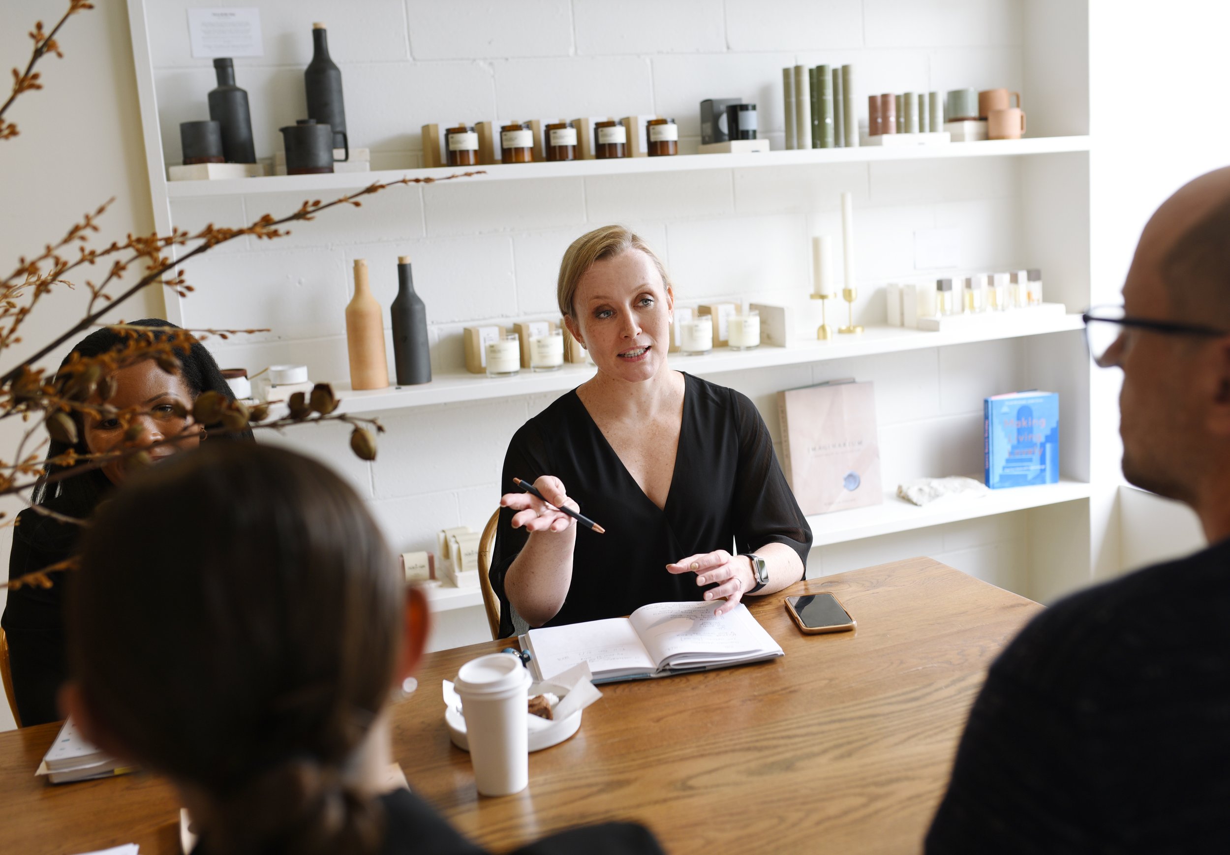 A woman with blonde hair speaking in a discussion group, seated at a wooden table with notebooks, a smartphone, and a disposable coffee cup. Several people are partially visible around her, with shelves containing candles, bottles, and mugs in the background.