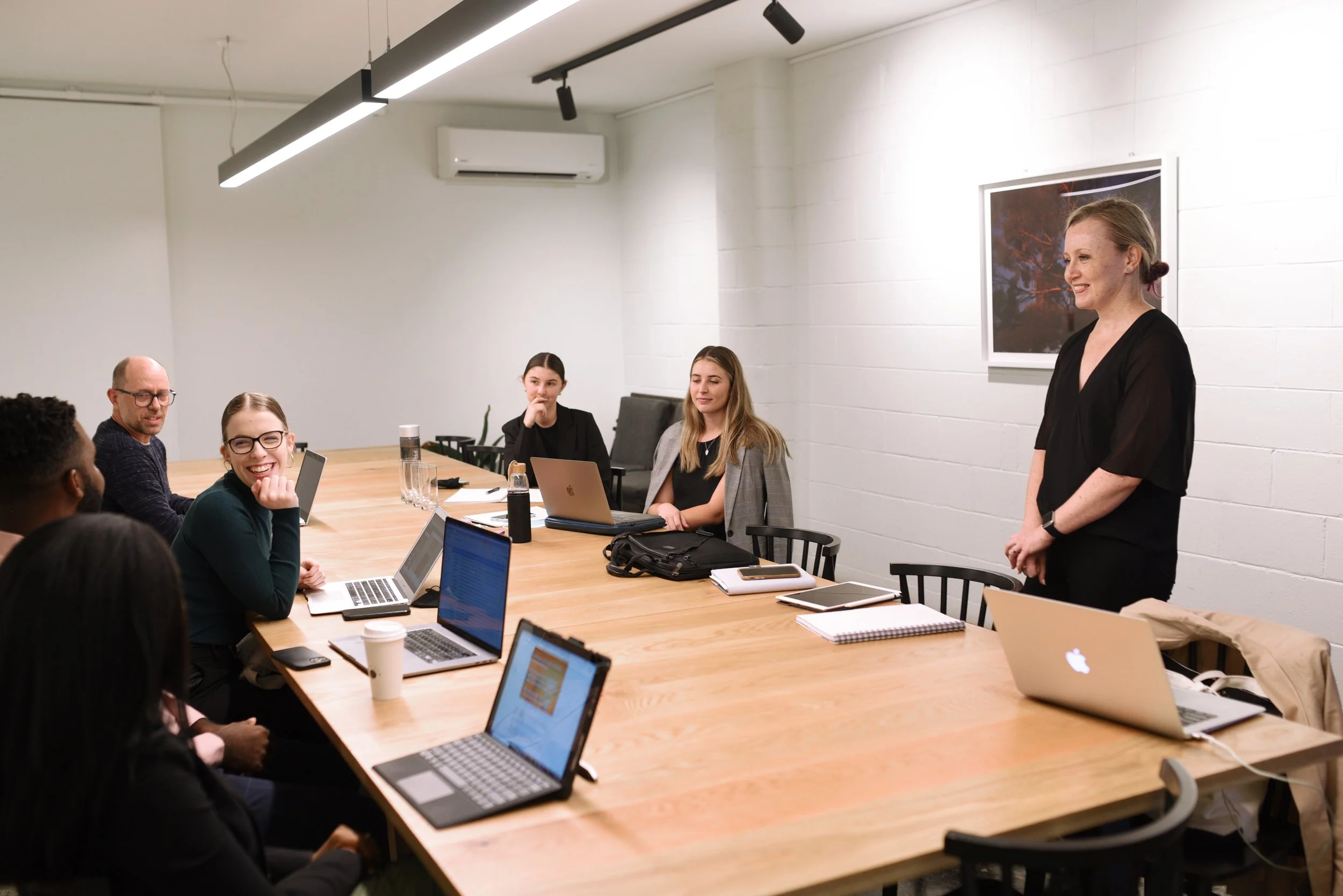 A woman is standing and speaking to a group of people seated around a long conference table, with laptops and notebooks in a modern, well-lit room.