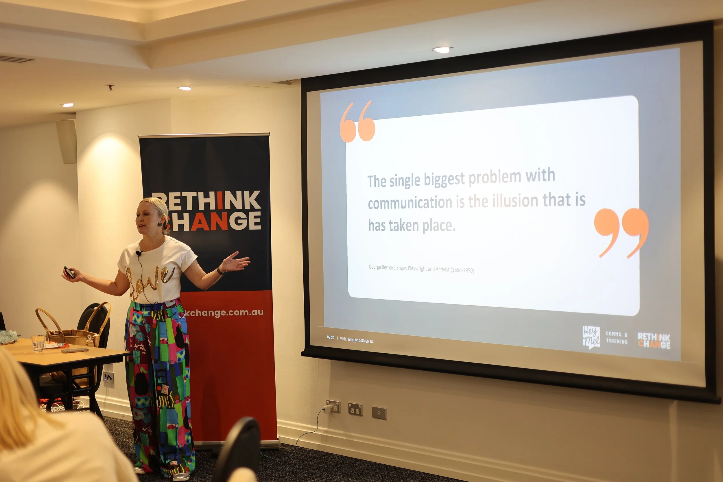 A woman giving a presentation in a conference room, with a Large screen displaying a quote by George Bernard Shaw about communication, and a banner behind her that reads 'RETHINK CHANGE'.