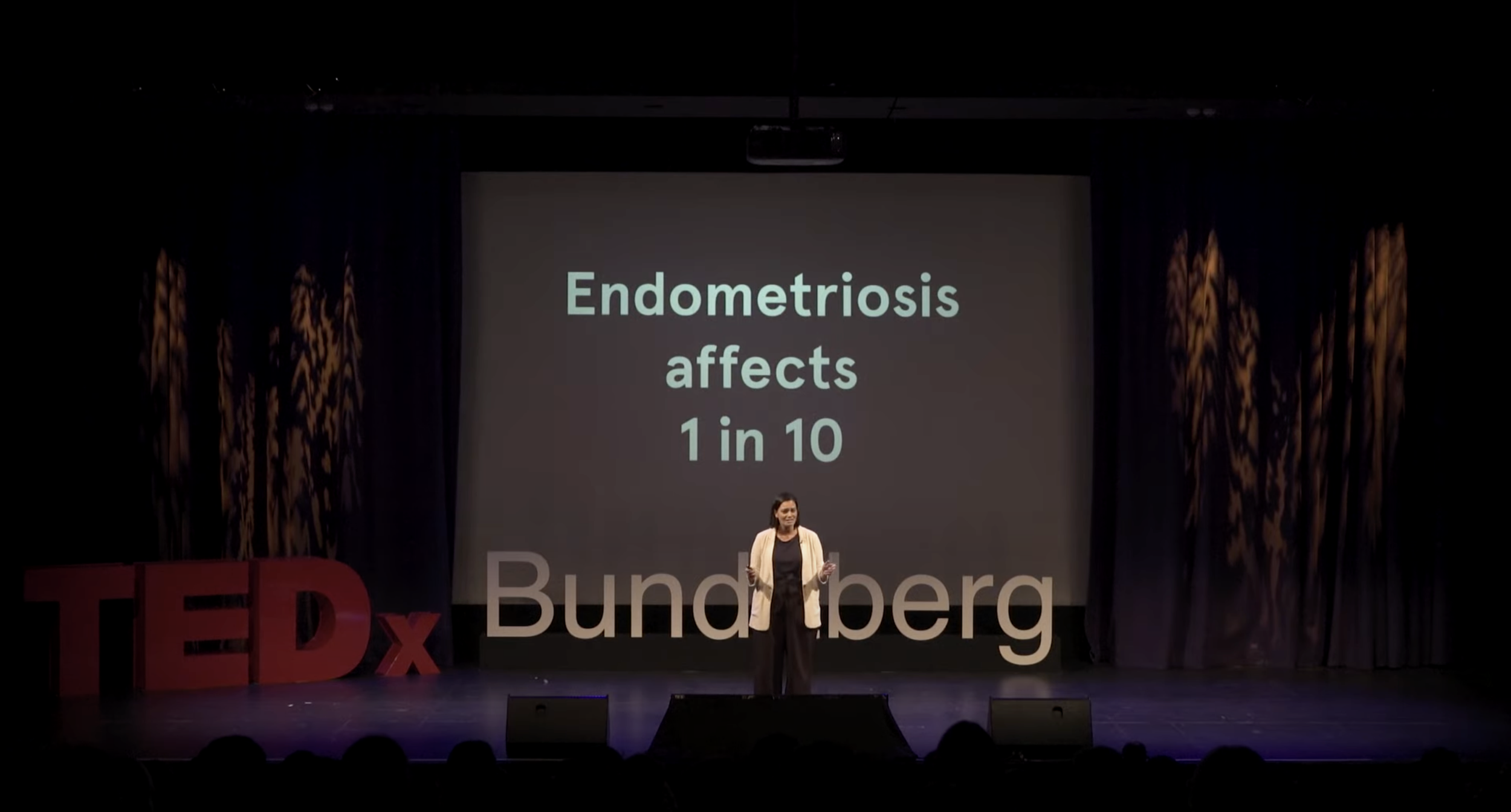 A woman stands on stage at a TEDx event, with a large screen behind her displaying the text 'Endometriosis affects 1 in 10.' Large red TEDx letters are on the left side of the stage.
