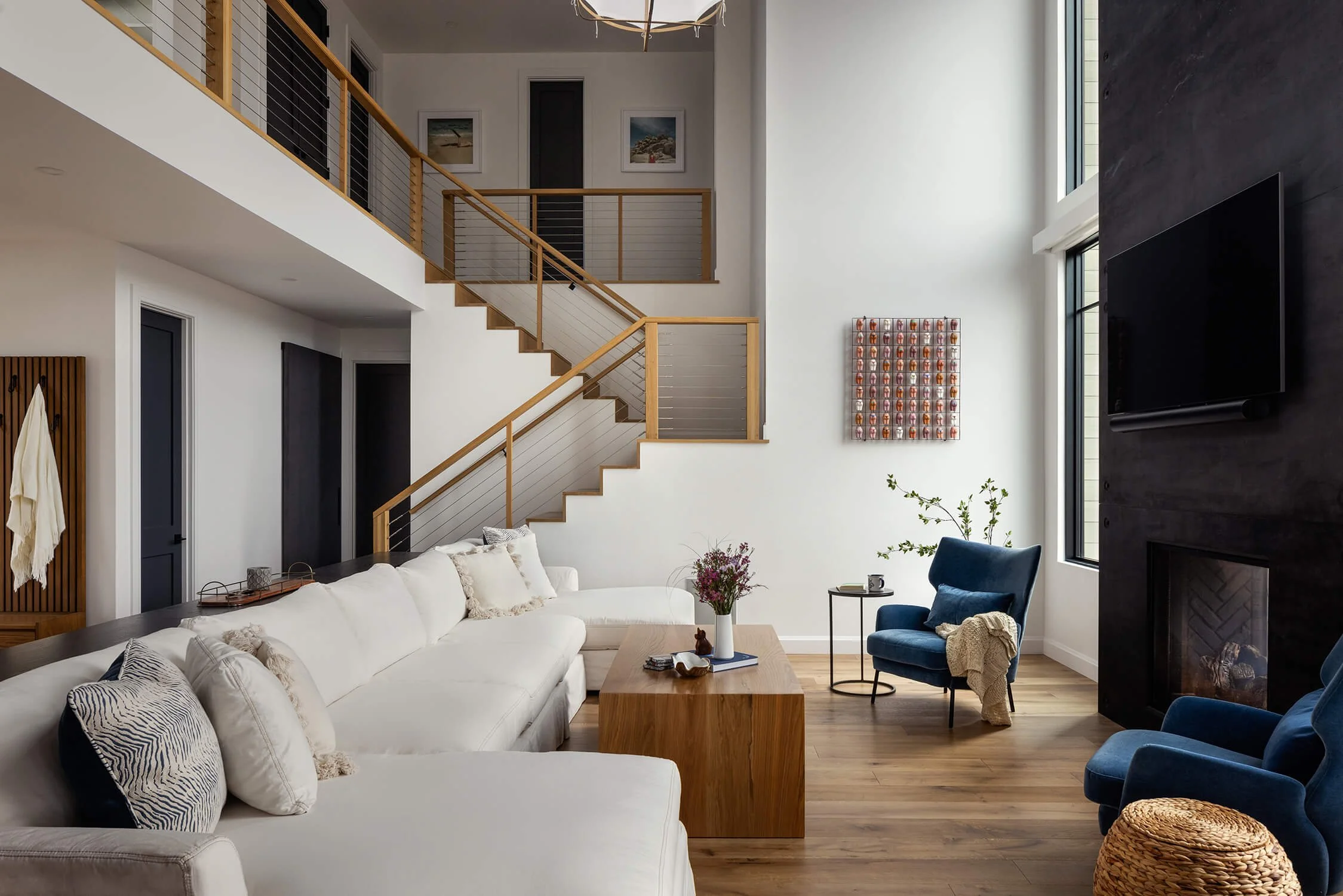 Double height living room with cable staircase, white sectional sofa, dark fireplace wall, blue velvet accent chairs, and abstract wall art, photographed by Brian Berkowitz Photography in New York