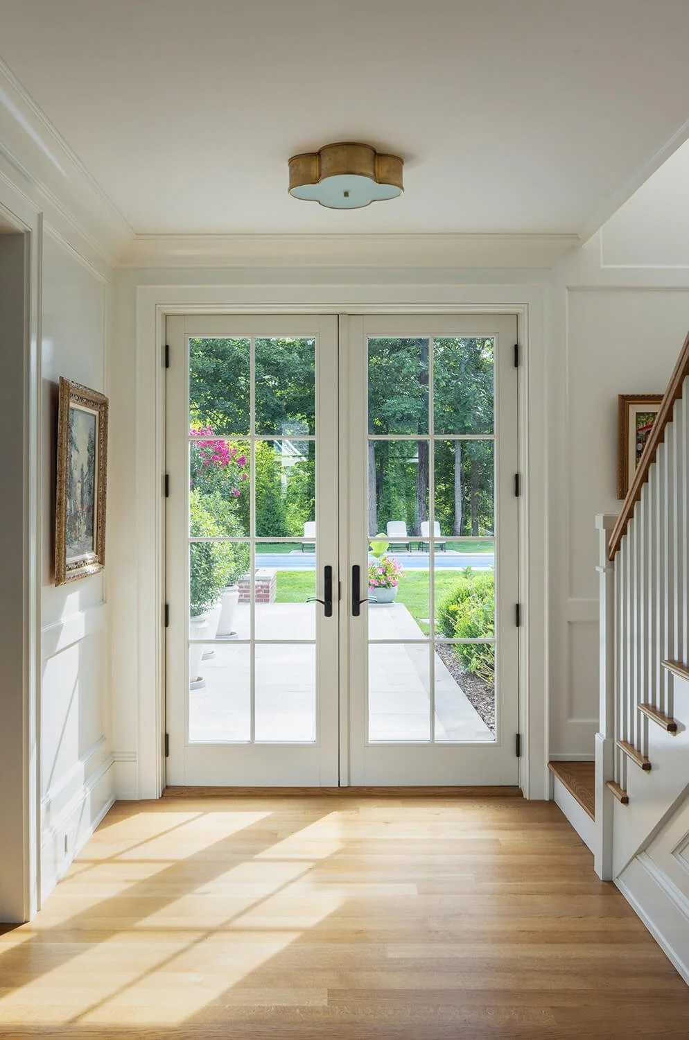 Bright entry foyer with white French doors opening to the garden, hardwood floors, staircase, and brass flush mount ceiling fixture, photographed by Brian Berkowitz Photography on Long Island