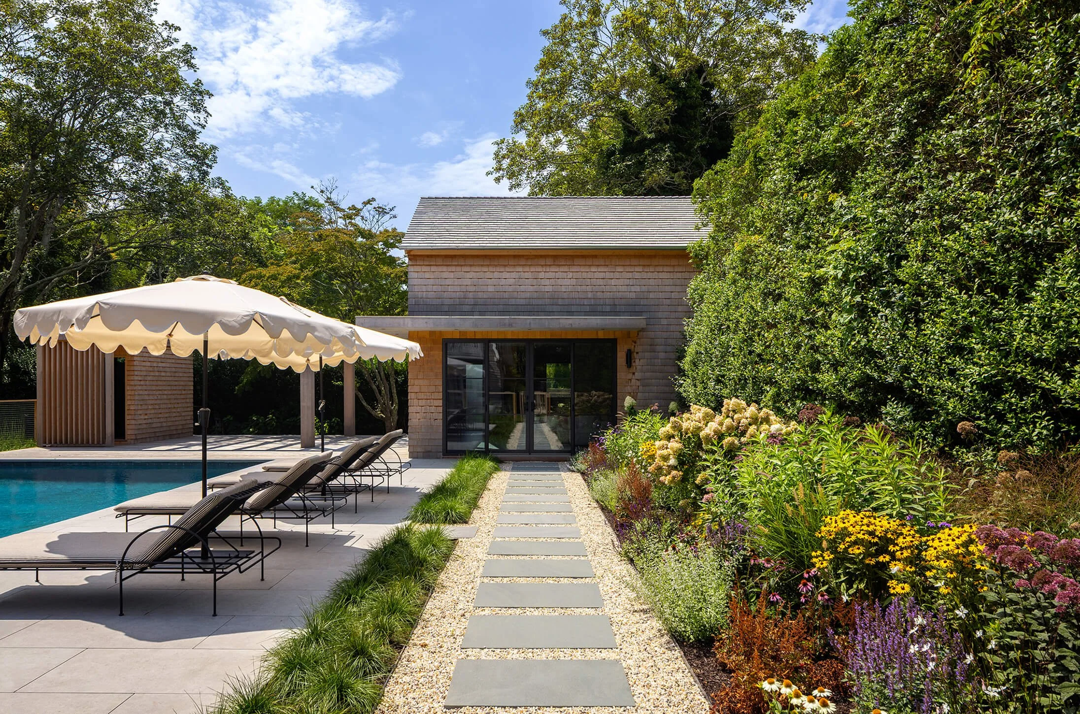 Hamptons pool house with cedar shingle exterior, stone garden path, scalloped market umbrella, lounge chairs, and lush perennial garden border, photographed by Brian Berkowitz Photography