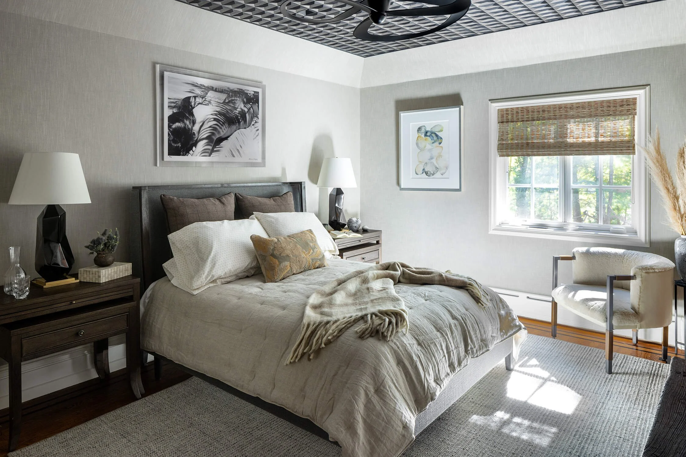 bedroom with geometric wallpapered ceiling, wood headboard, layered bedding, sculptural lamp, and black and white photography above the bed, photographed by Brian Berkowitz Photography on Long Island