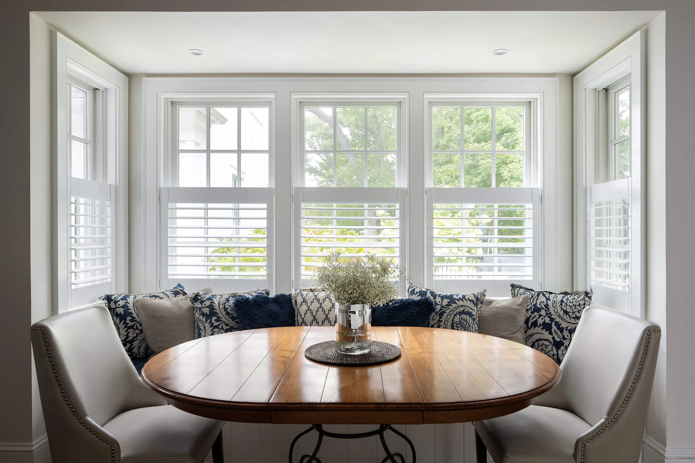 Breakfast nook with banquette seating, navy and white patterned pillows, round wood dining table, and bay window with plantation shutters, photographed by Brian Berkowitz Photography on Long Island