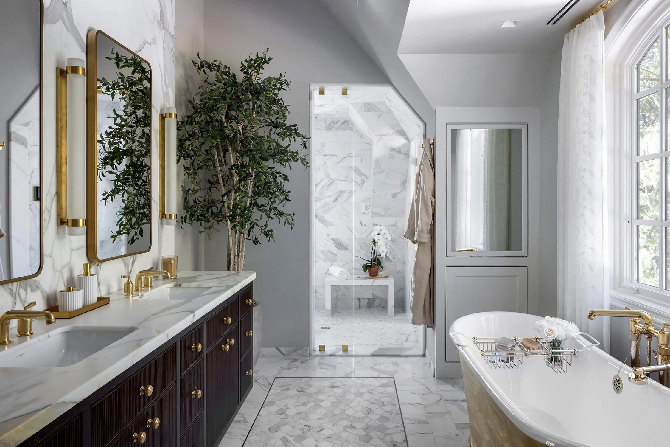 Primary bathroom with Calacatta marble walls and floors, brass fixtures, double vanity, freestanding soaking tub, and indoor olive tree, photographed by Brian Berkowitz Photography on Long Island
