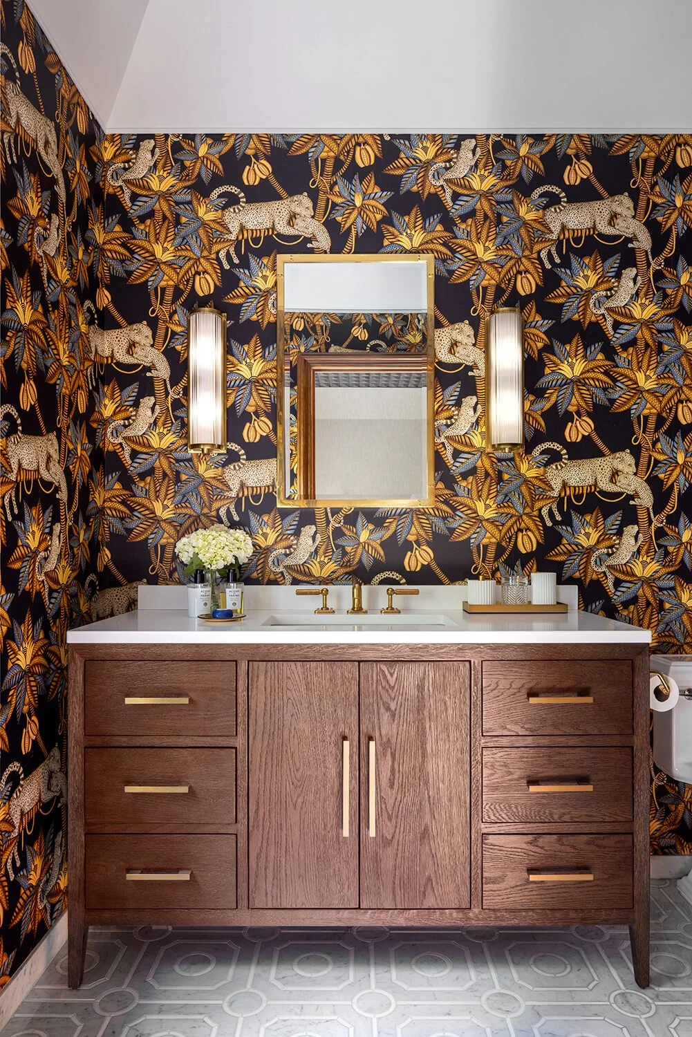 Powder room with leopard and floral wallpaper, dark wood vanity with brass hardware, gold framed mirror, and geometric cement tile floor, photographed by Brian Berkowitz Photography on Long Island