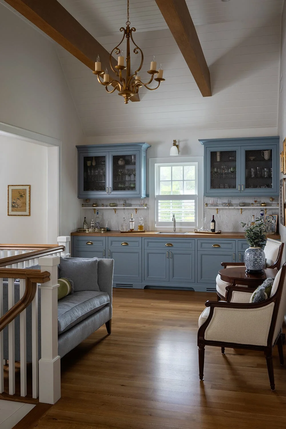 Bar room and sitting area with blue cabinetry, brass hardware, shiplap ceiling, exposed wood beams, brass chandelier, and staircase railing, photographed by Brian Berkowitz Photography on Long Island