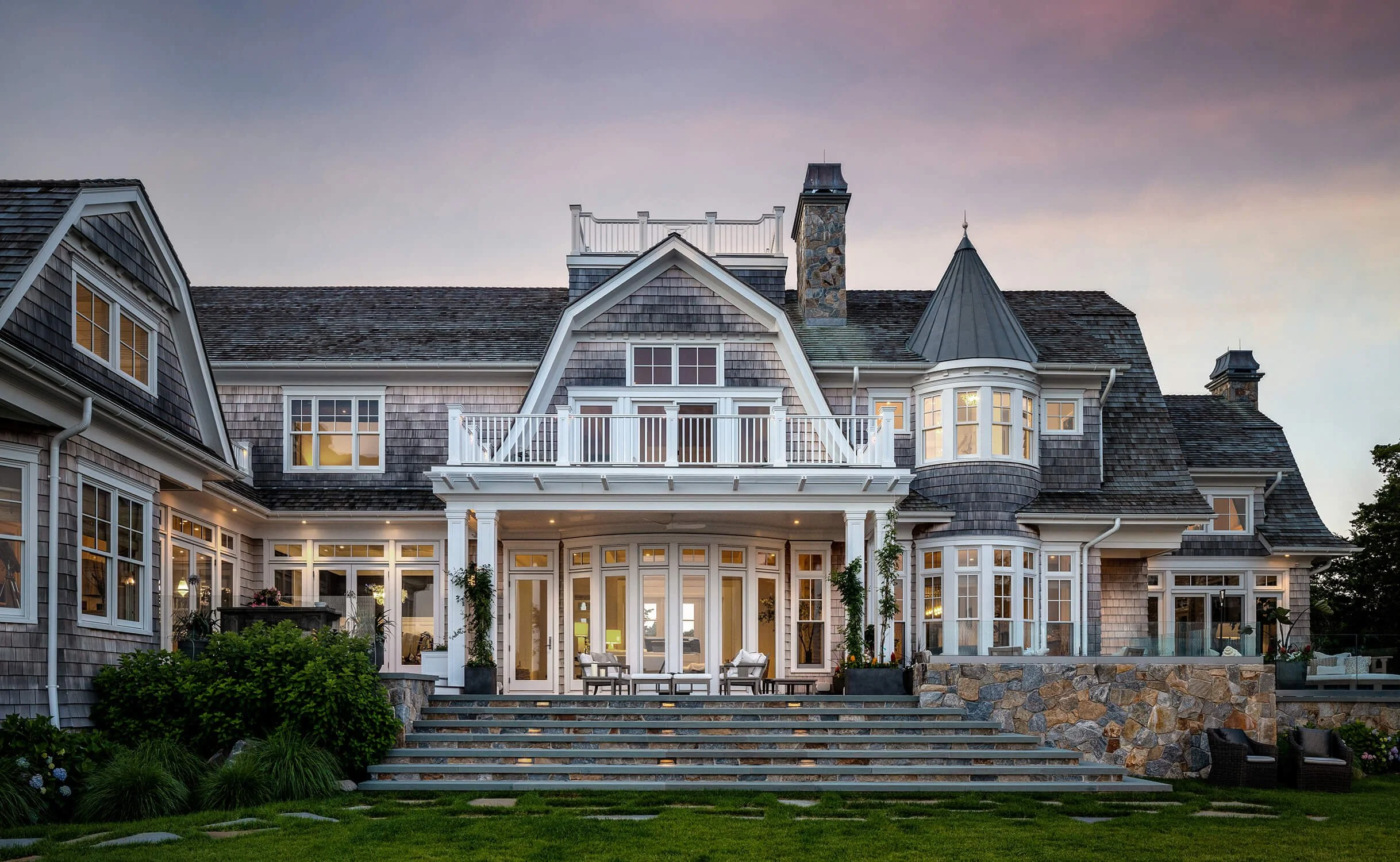 Grand Hamptons style shingle mansion at dusk with illuminated windows, stone staircase, manicured landscaping, and dramatic purple sky, photographed by Brian Berkowitz Photography on Long Island