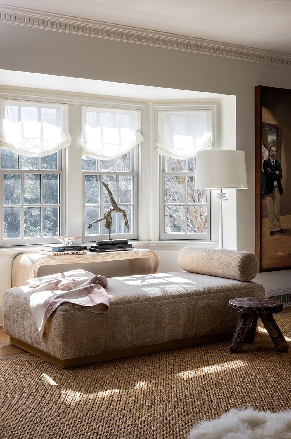 Sitting room with upholstered daybed, bay window with roman shades, bronze sculpture, large-scale portrait painting, and natural light, photographed by Brian Berkowitz Photography on Long Island
