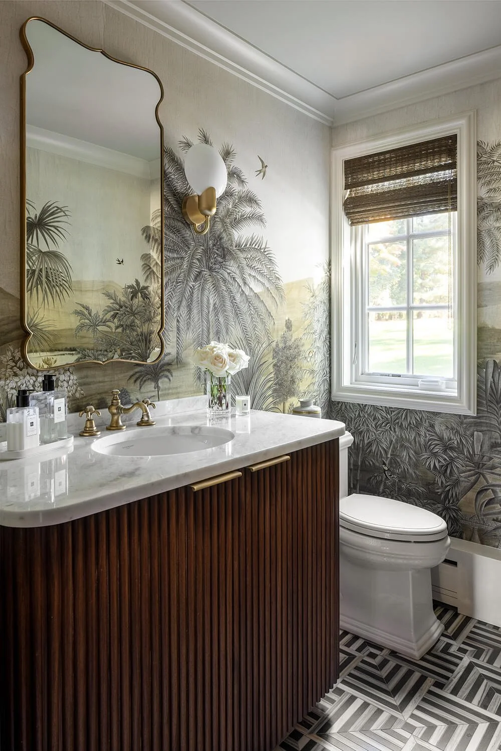 Powder room with scenic tropical mural wallpaper, fluted dark wood vanity, marble countertop, ornate brass mirror, and geometric floor tile, photographed by Brian Berkowitz Photography on Long Island