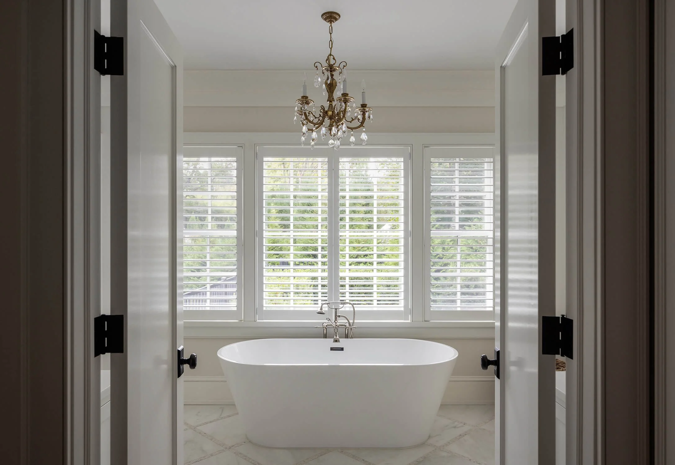 bathroom with soaking tub centered beneath a chandelier, floor-to-ceiling plantation shutters, marble tile floor, and framed doorway entry, photographed by Brian Berkowitz Photography on Long Island