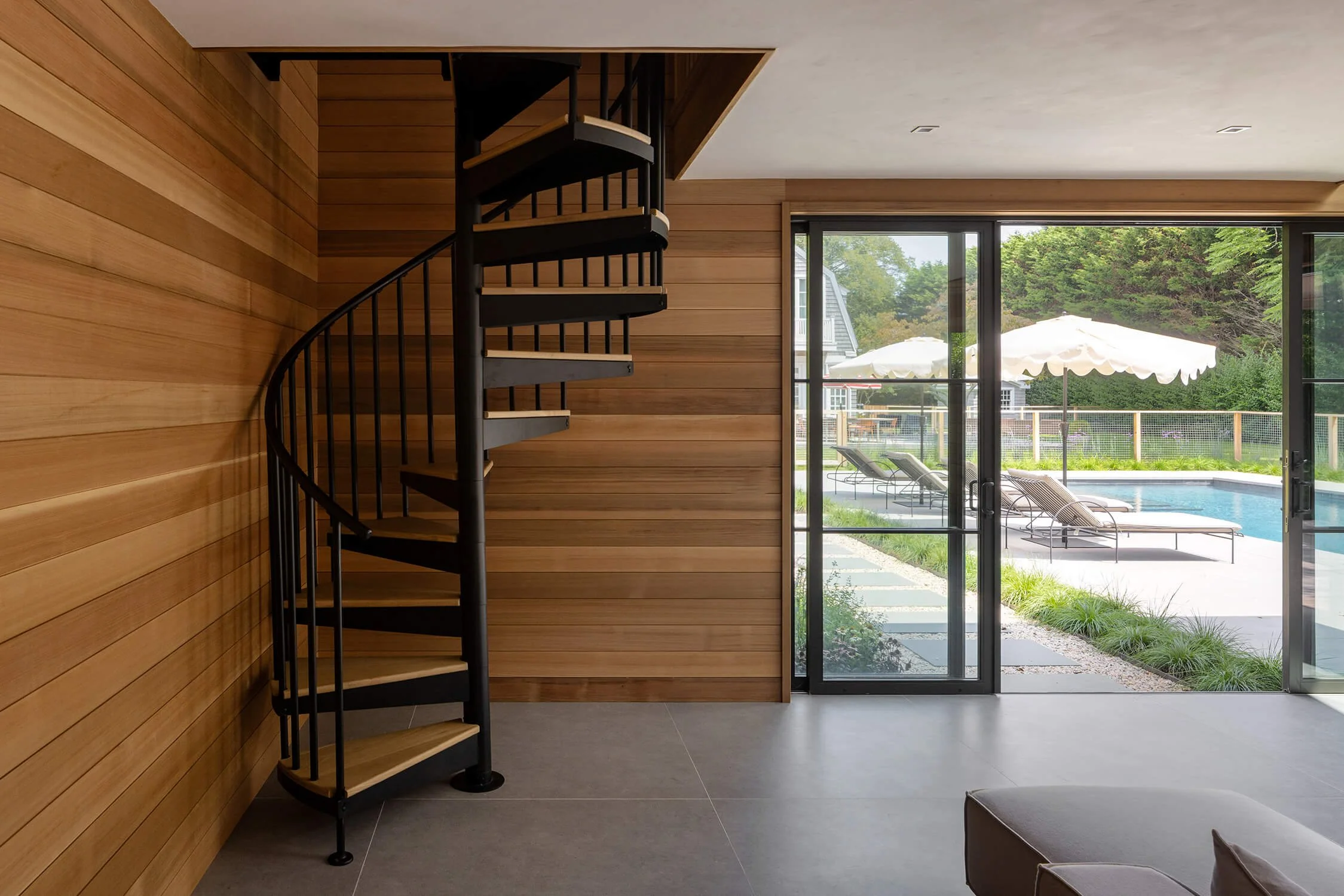 Pool house interior with black spiral staircase, cedar shiplap walls, concrete tile floor, and sliding doors opening to the pool and garden, photographed by Brian Berkowitz Photography in the Hamptons